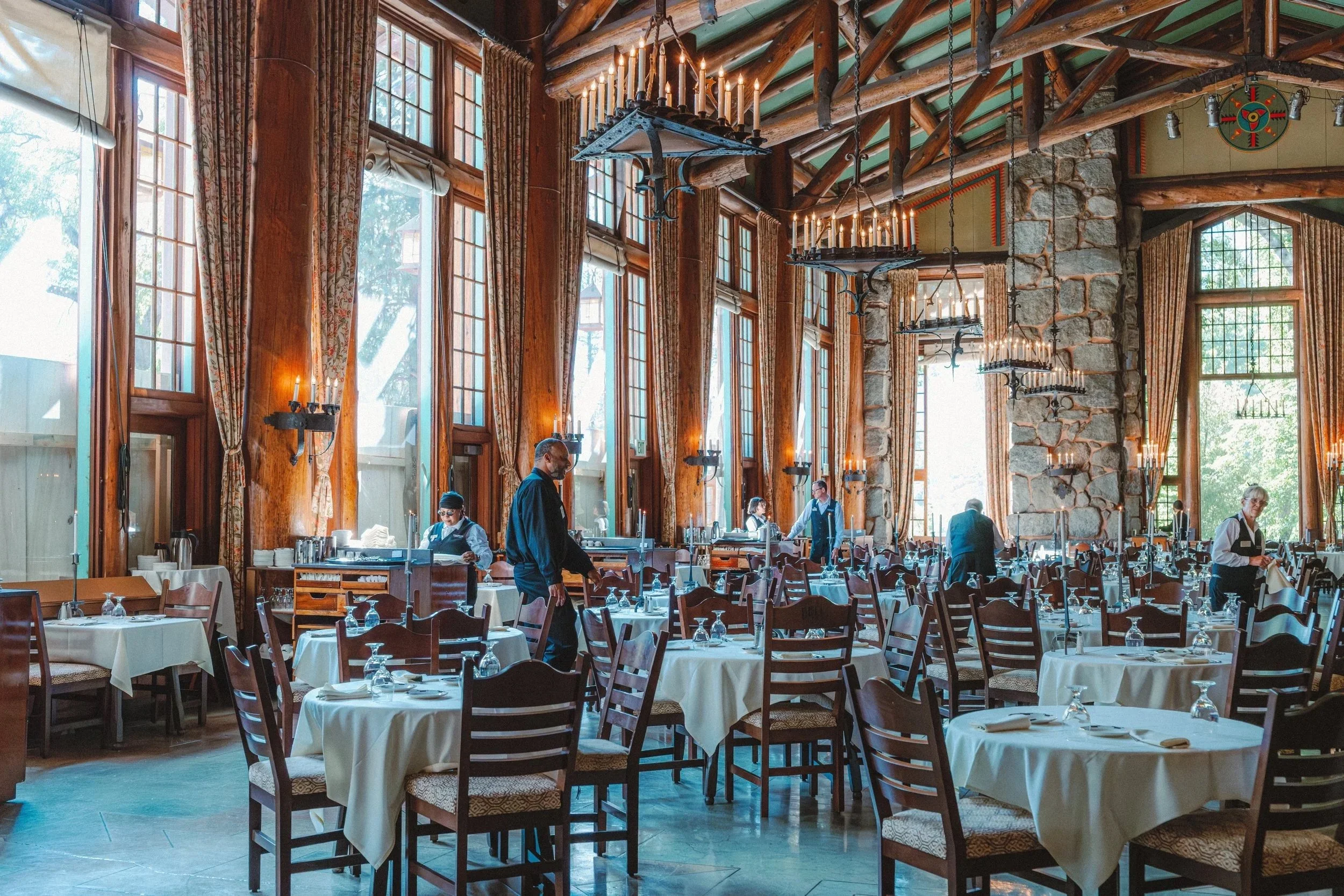 Elegant restaurant interior with tall windows, wooden beams, stone accents, and chandeliers, set with white tablecloths and chairs, and staff preparing tables.