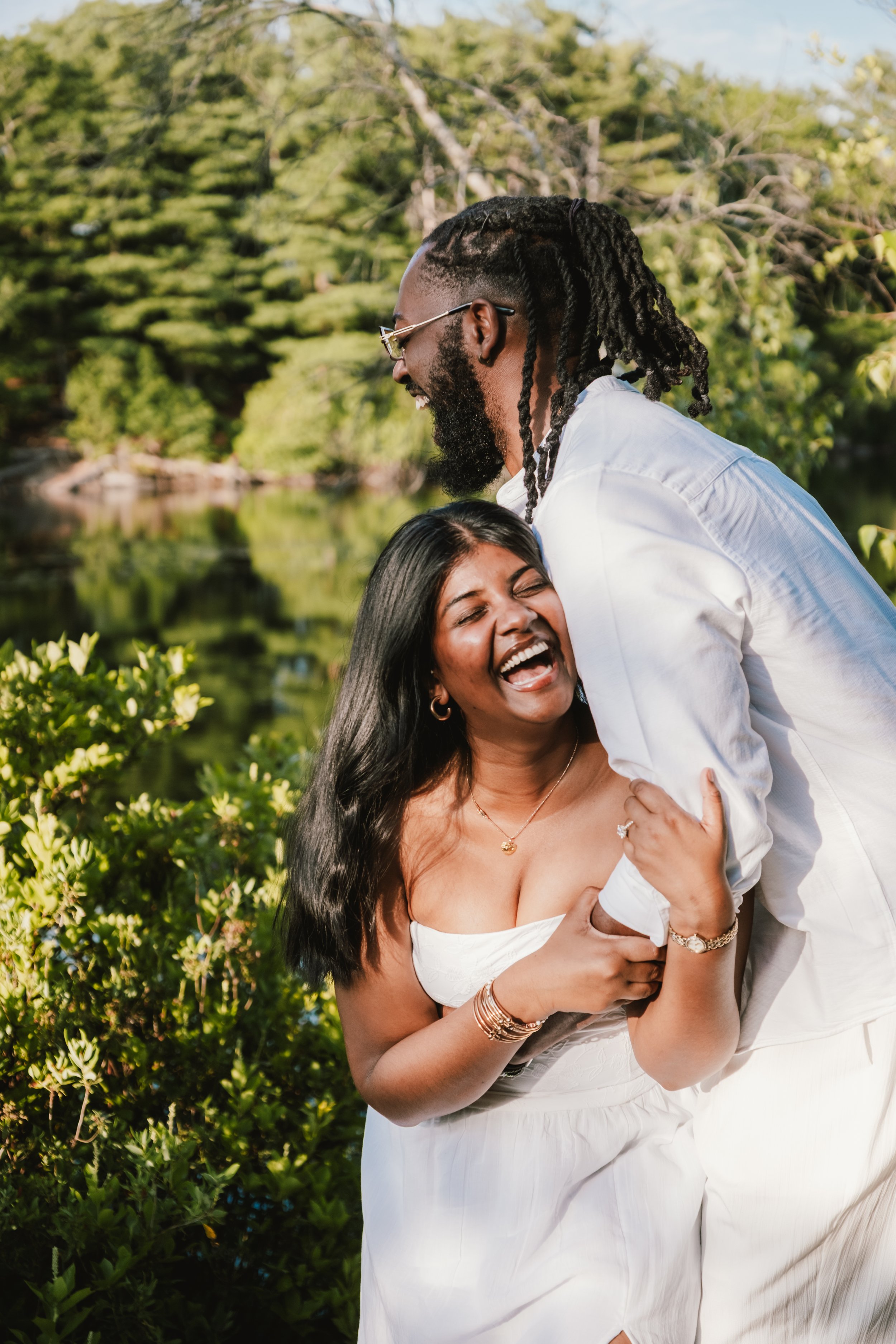A joyful couple is laughing and hugging outdoors near a body of water with trees in the background.