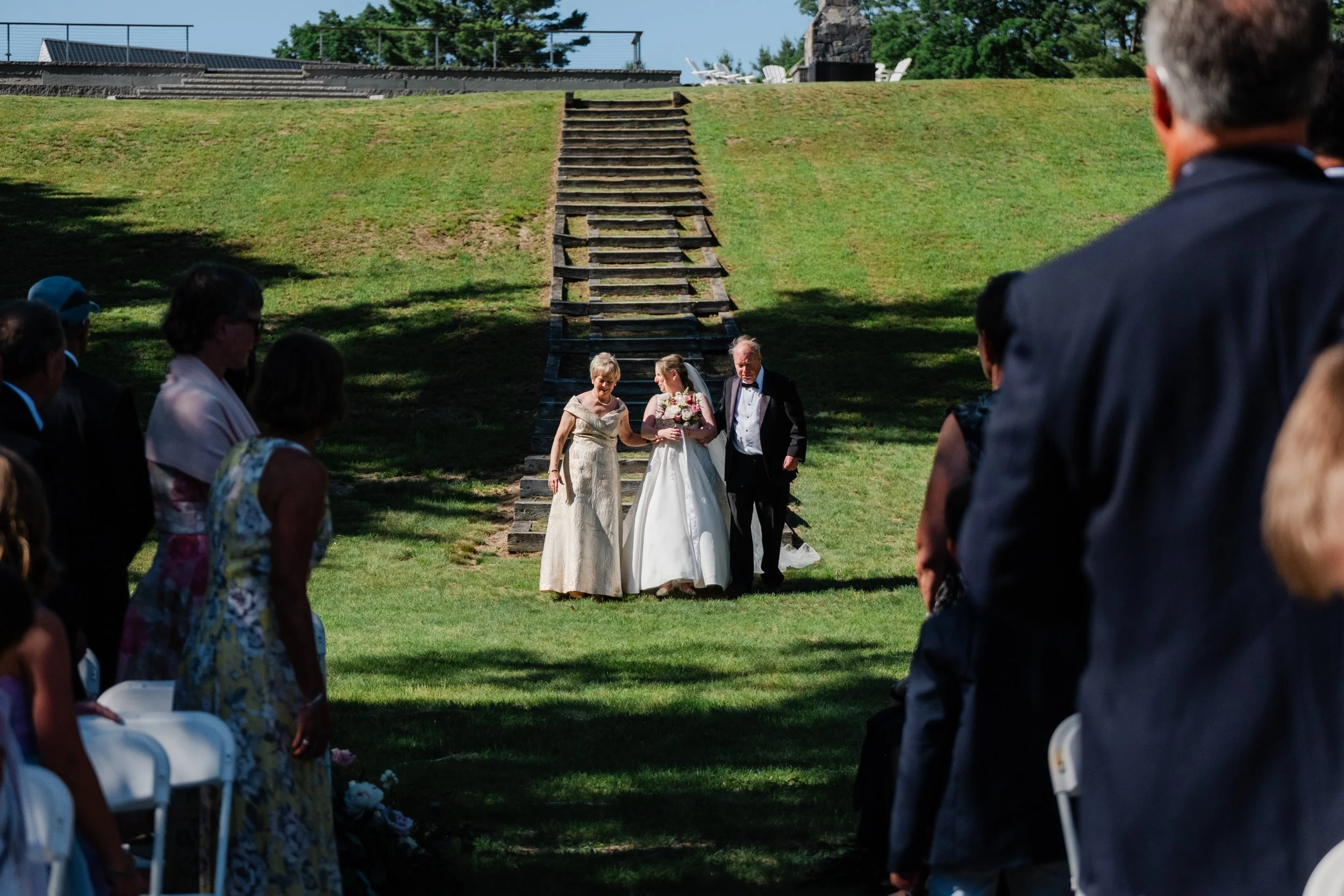 A bride is escorted by her parents down a set of stairs at Bear Mountain Inn + Barn during her Maine wedding.