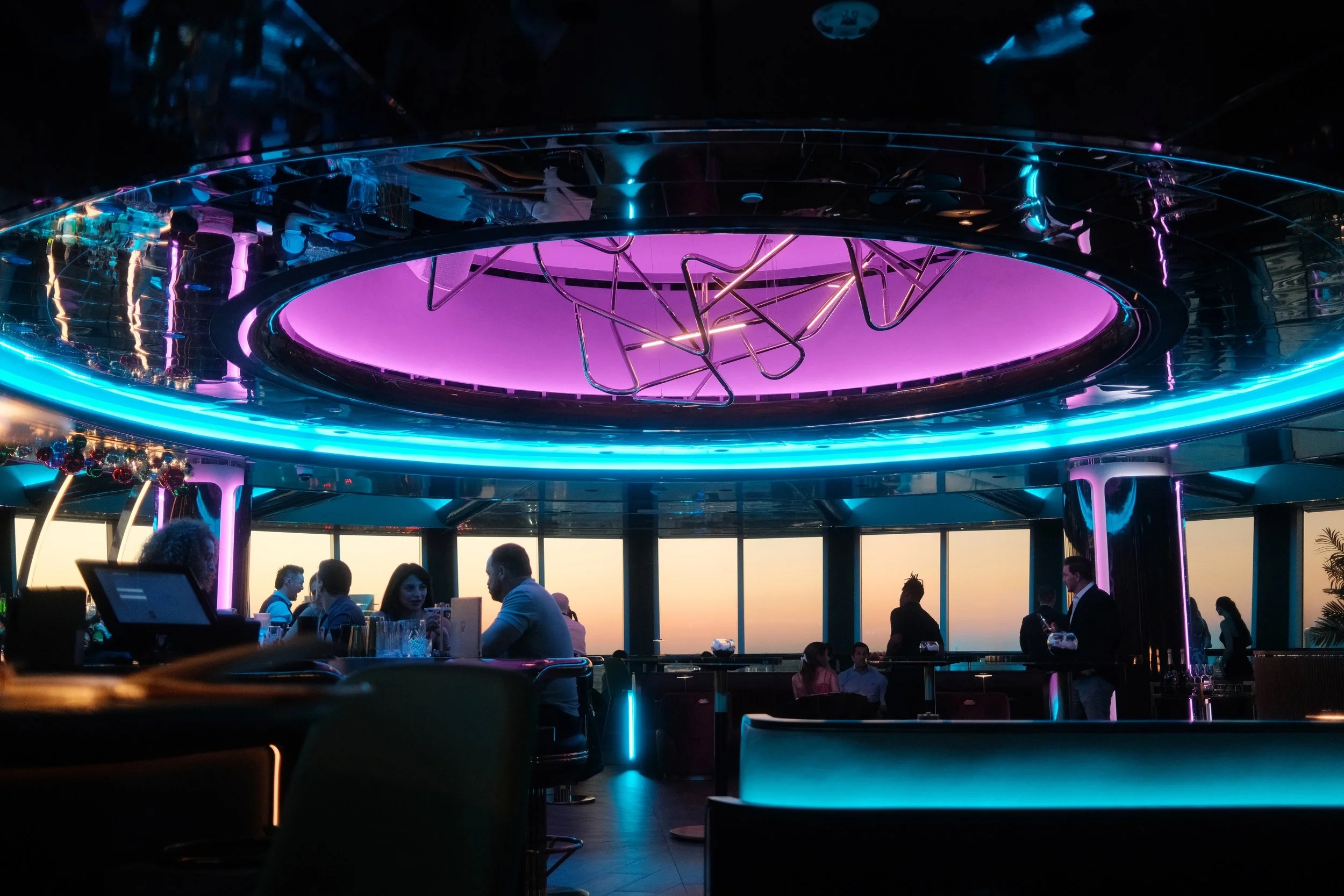 Interior of a modern rotating rooftop bar restaurant at Pier Sixty-Six in Fort Lauderdale, Florida with colorful neon lighting, large windows showing a sunset sky, and people sitting at tables and standing, possibly ordering drinks.