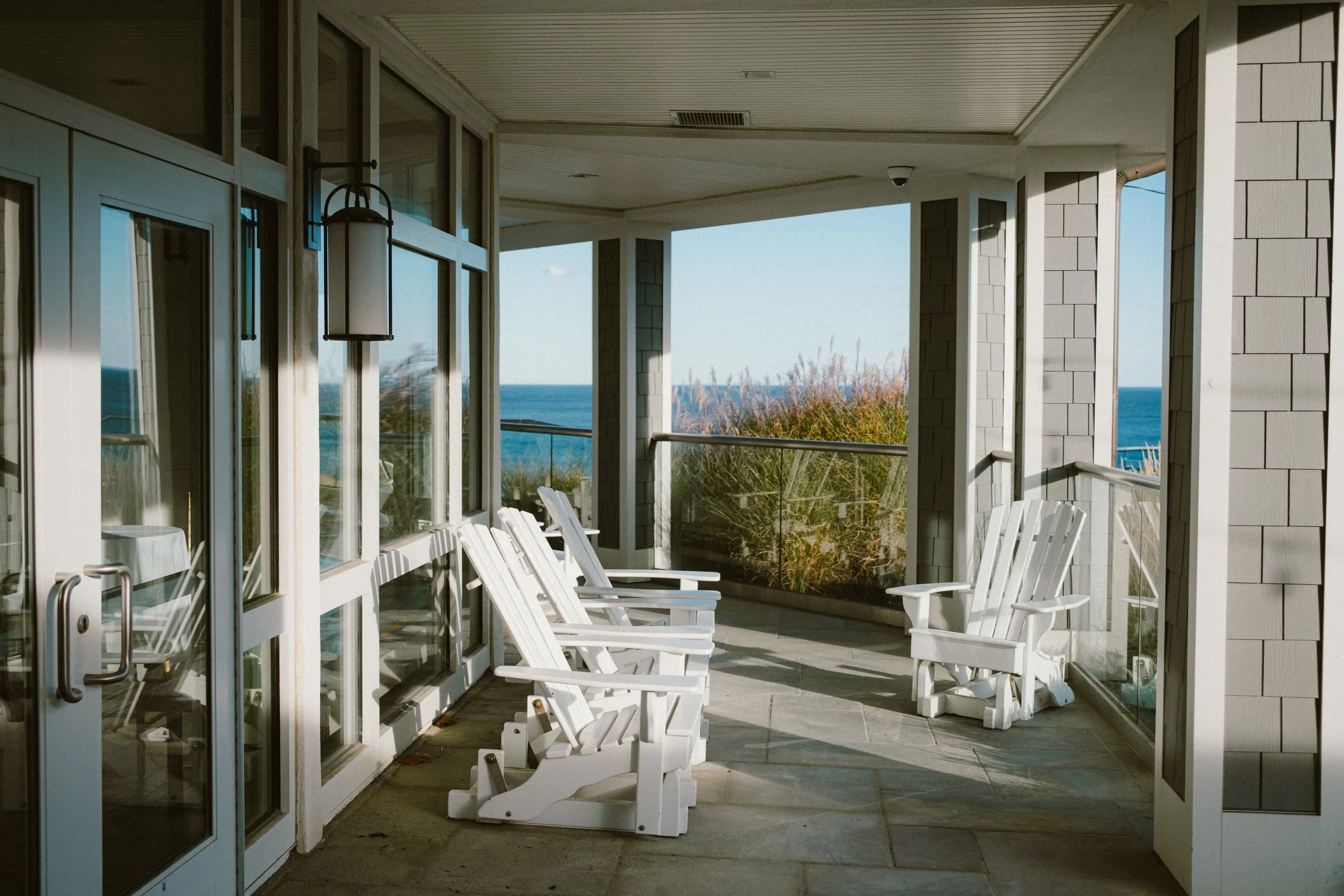 A balcony with white Adirondack chairs overlooking the ocean, featuring glass railing and a view of blue water and sky at the Cliff House Hotel in Maine.