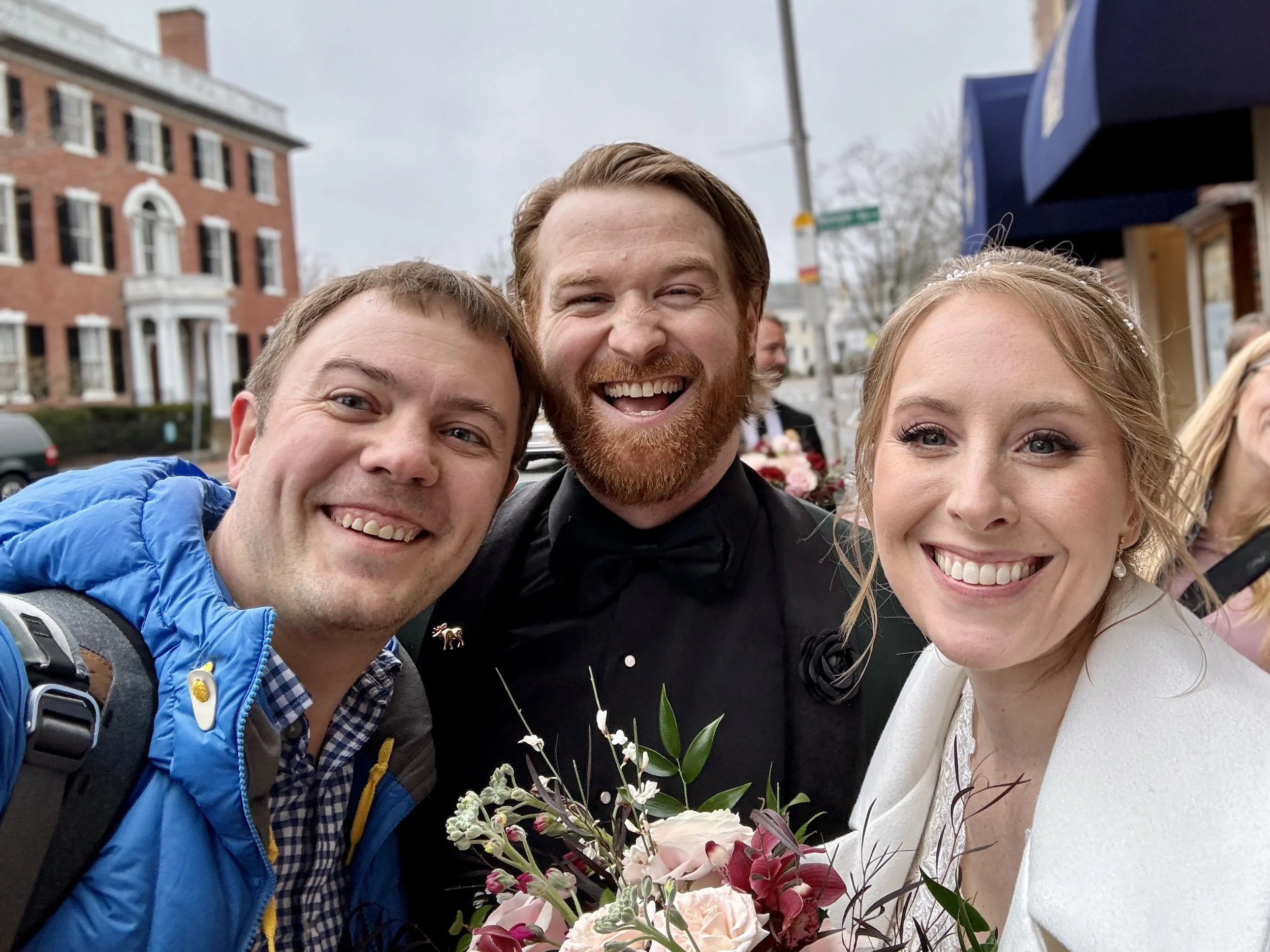 Three smiling people, two men and one woman, taking a selfie outdoors near a wedding or celebration. The woman holds a bouquet of flowers.