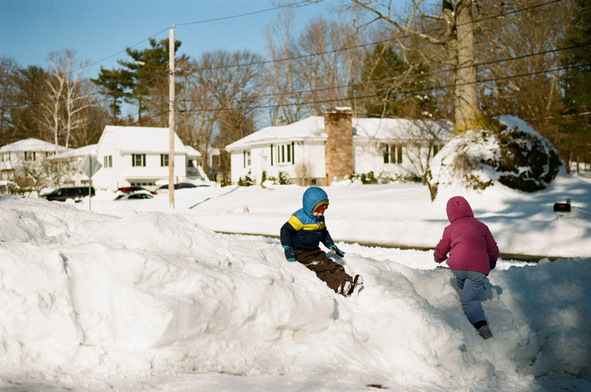 Two children playing in the snow on a suburban street, climbing a snow mound. One child is wearing a blue and yellow jacket with a blue hood, and the other is in a pink jacket with a hood. Snow-covered trees, houses, and a clear blue sky are visible in the background.