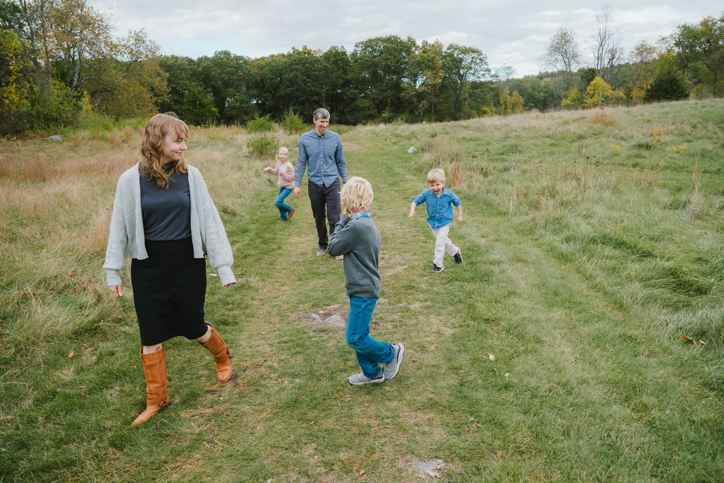 A family of six enjoying time together outdoors on a grassy trail in a rural area with trees in the background. The children are playing and running around while the adults watch and smile.