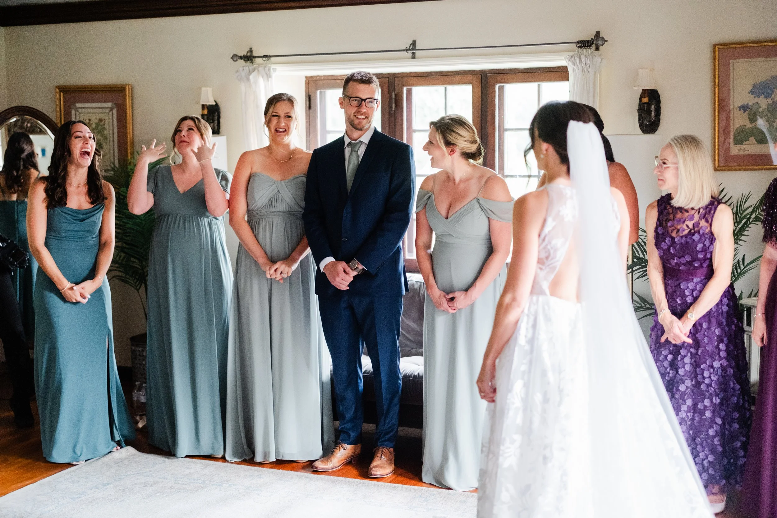 A wedding ceremony indoors with a bride facing a groom and a group of women in dresses watching and smiling.