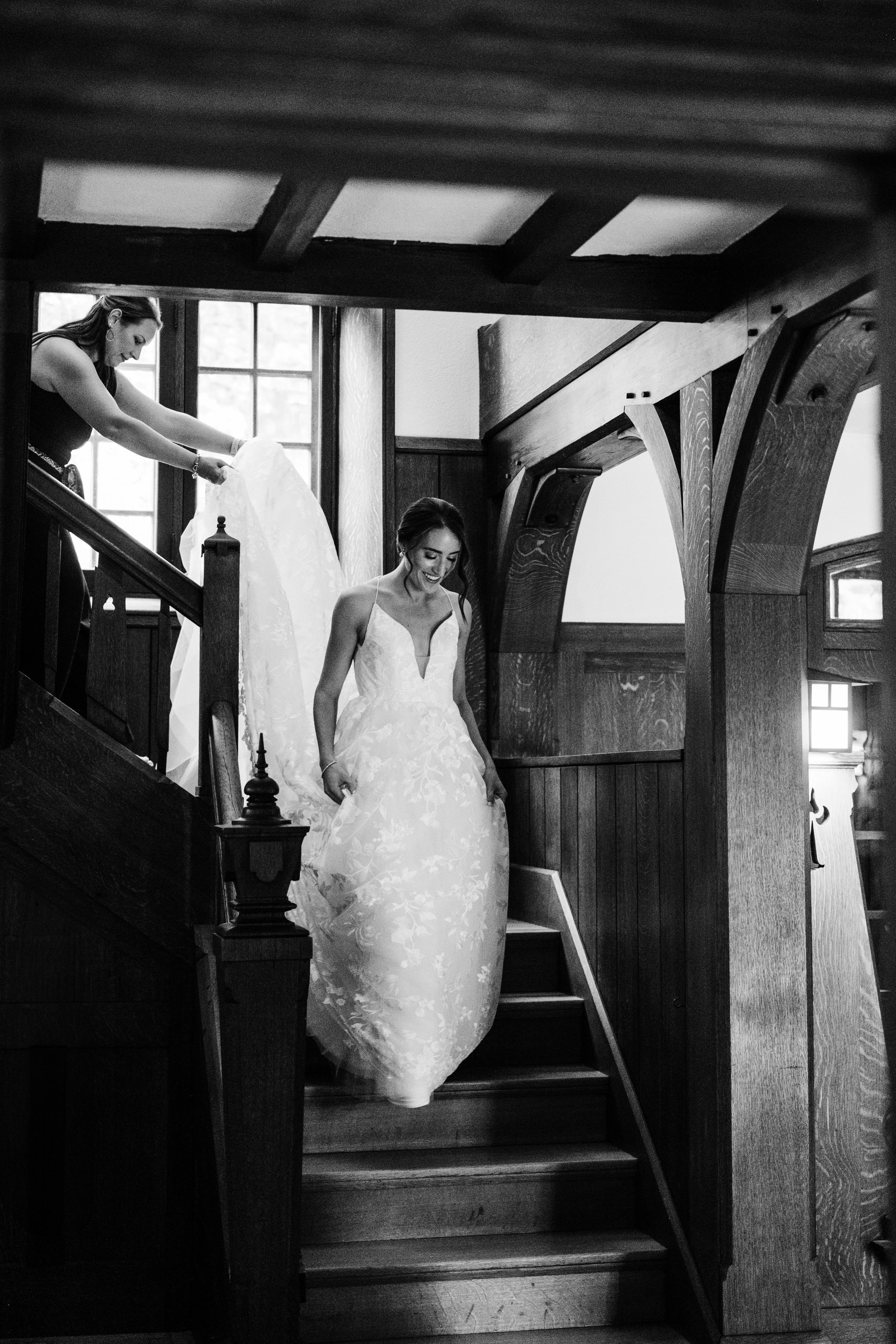 Bridal bride descending staircase in a vintage building with wood-paneled walls, assisted by a woman holding up her wedding dress.