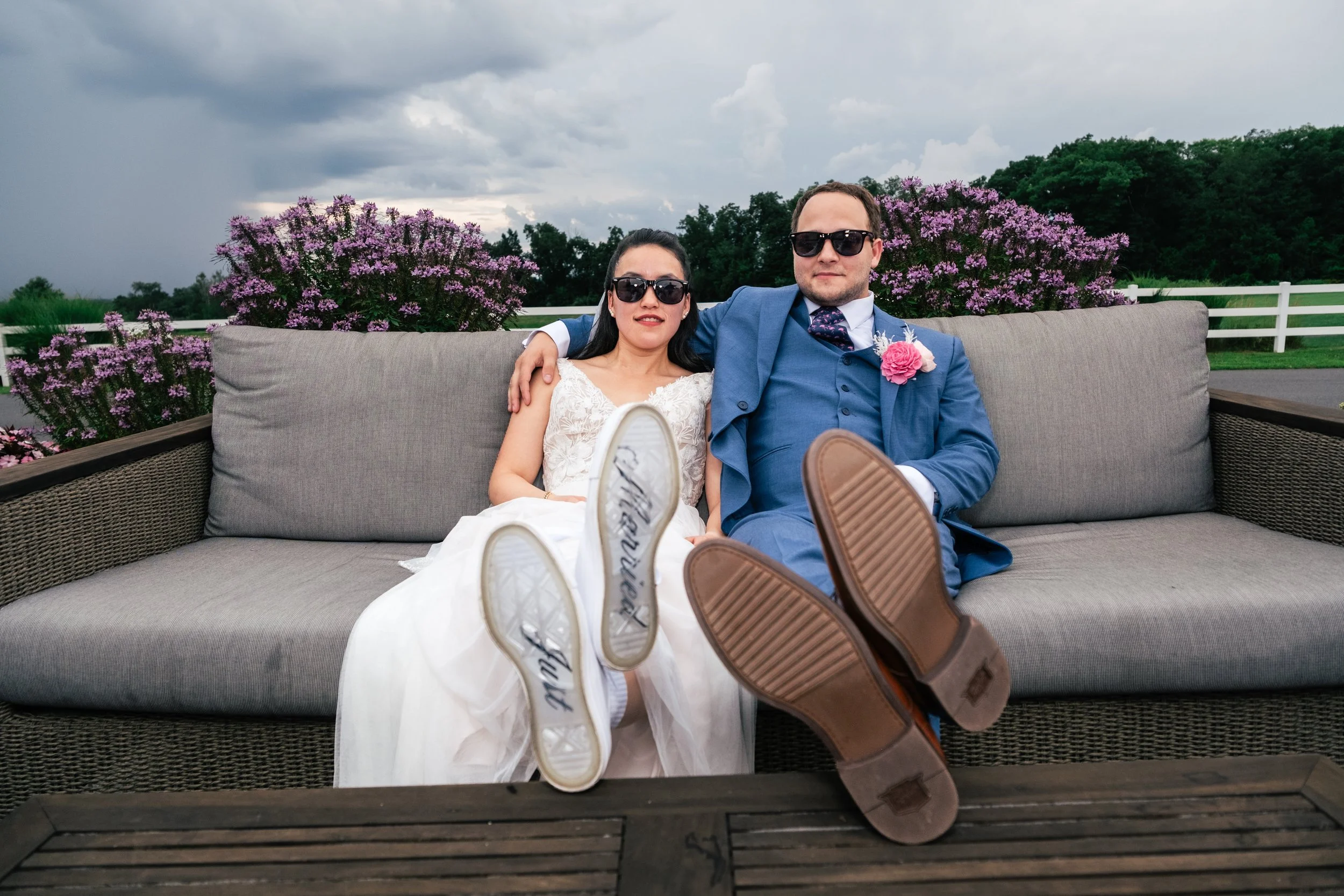 A bride and groom wear sunglasses while posing for a photo during their wedding at Four Oaks Country Club in Dracut, MA. A storm rolls in in the background and the bride wears custom sneakers that say "Just Married" on the bottom of the shoes.