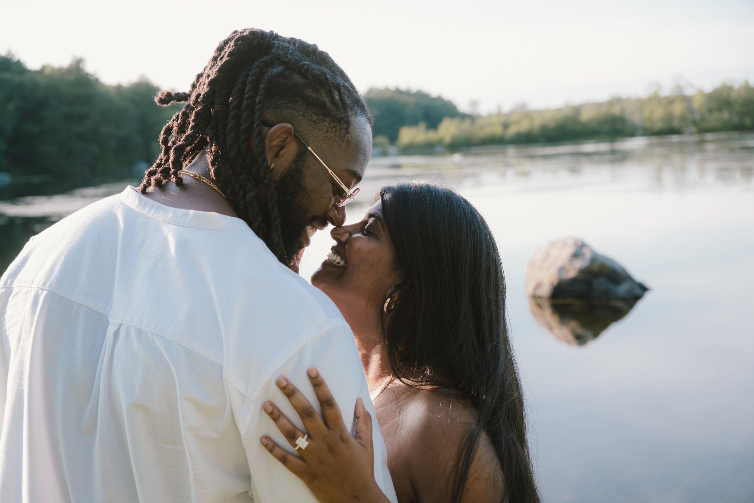 A couple smiling and embracing near a lake with a rock in the water, outdoors during daylight.