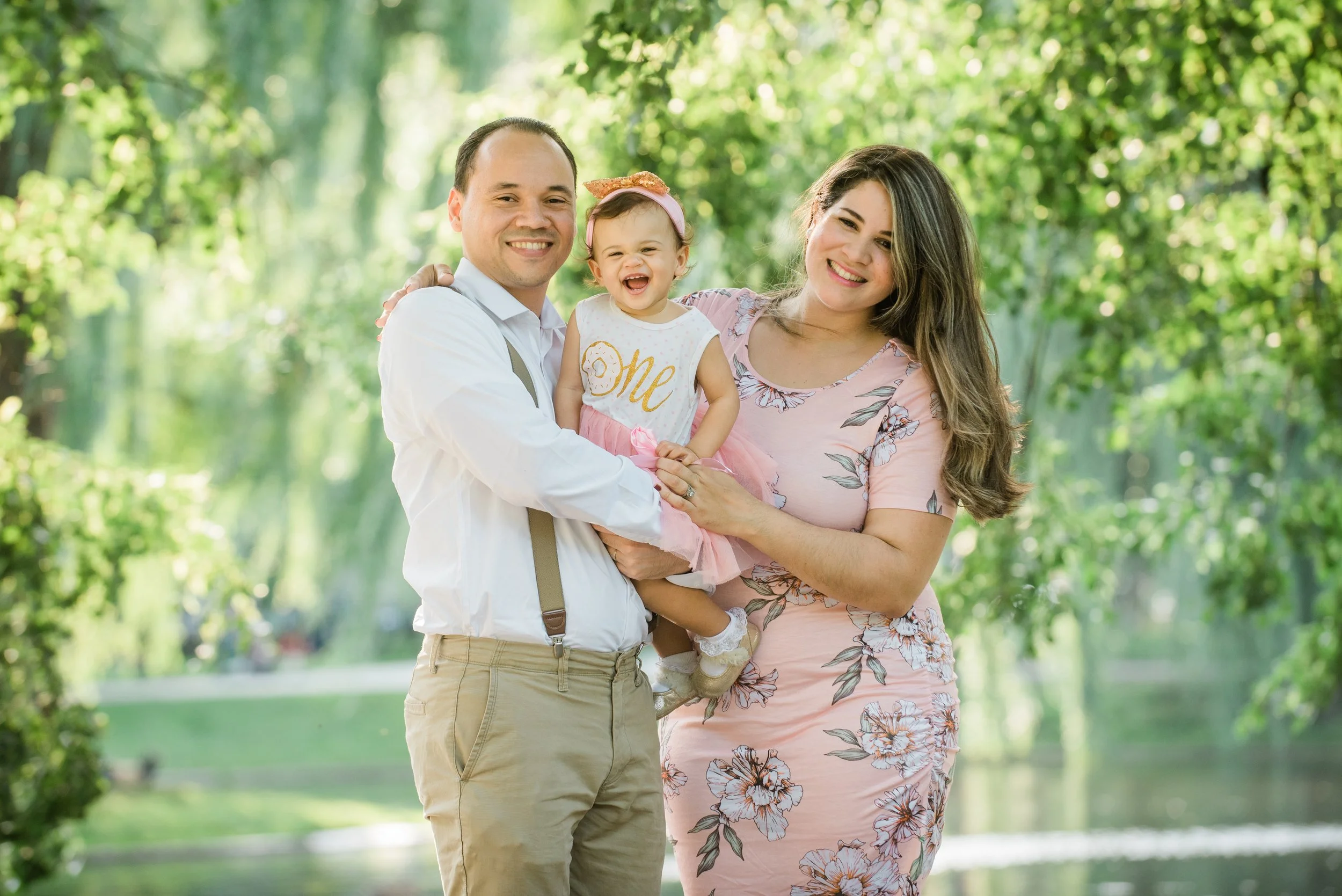 Smiling family of three, a man, woman, and young girl, outdoors in a lush green park with trees and a pond in the background. The man holds the girl, who wears a white shirt with the word "One" on it, and the woman stands beside them wearing a pink floral dress.