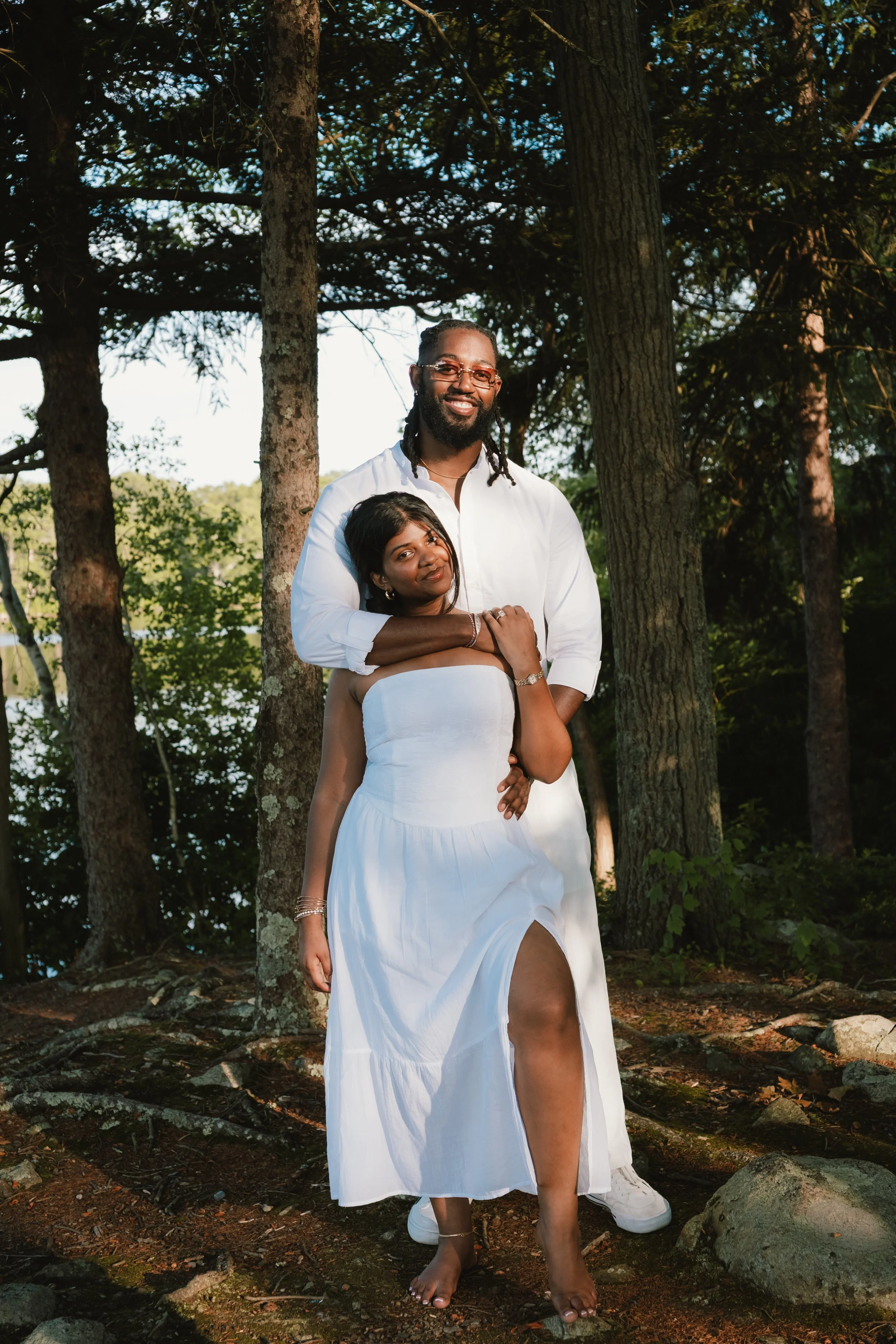 A smiling couple standing in a wooded area, with the woman in a strapless white dress and the man in a white shirt and pants, surrounded by trees and natural outdoor scenery.