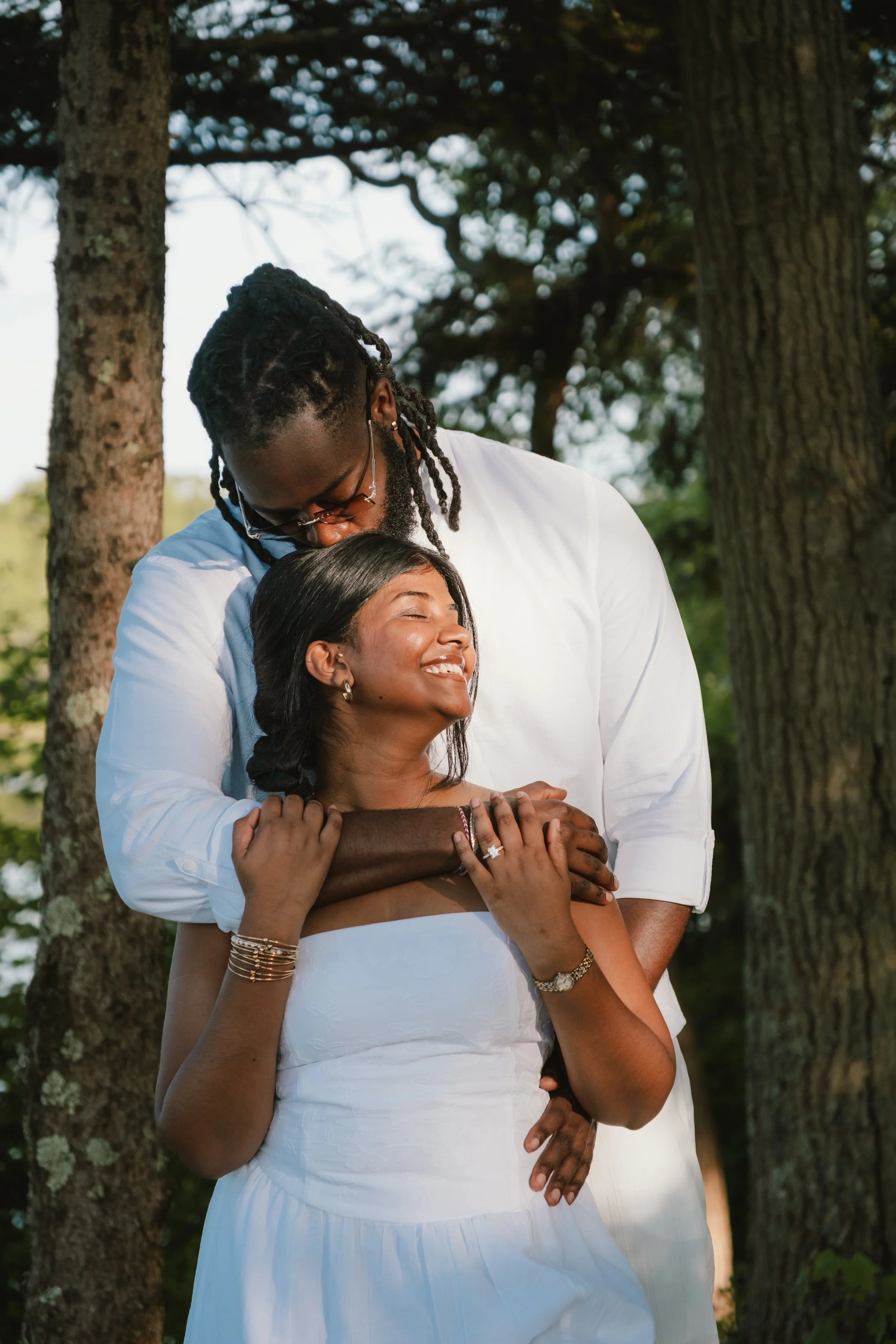 A happy couple embraces outdoors with trees in the background. The woman is smiling and has her eyes closed. The man is leaning over her, with his arms around her.