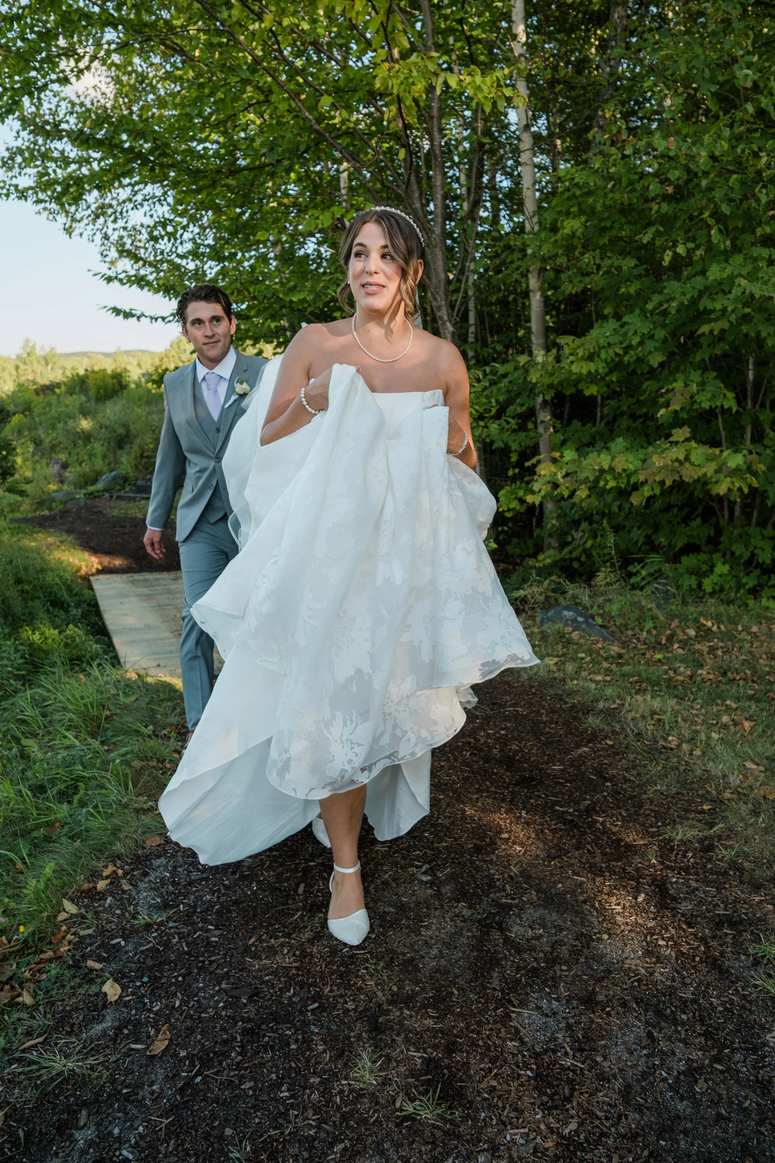 Bride in a white wedding gown walking outdoors with groom in a grey suit behind her on a wooded path