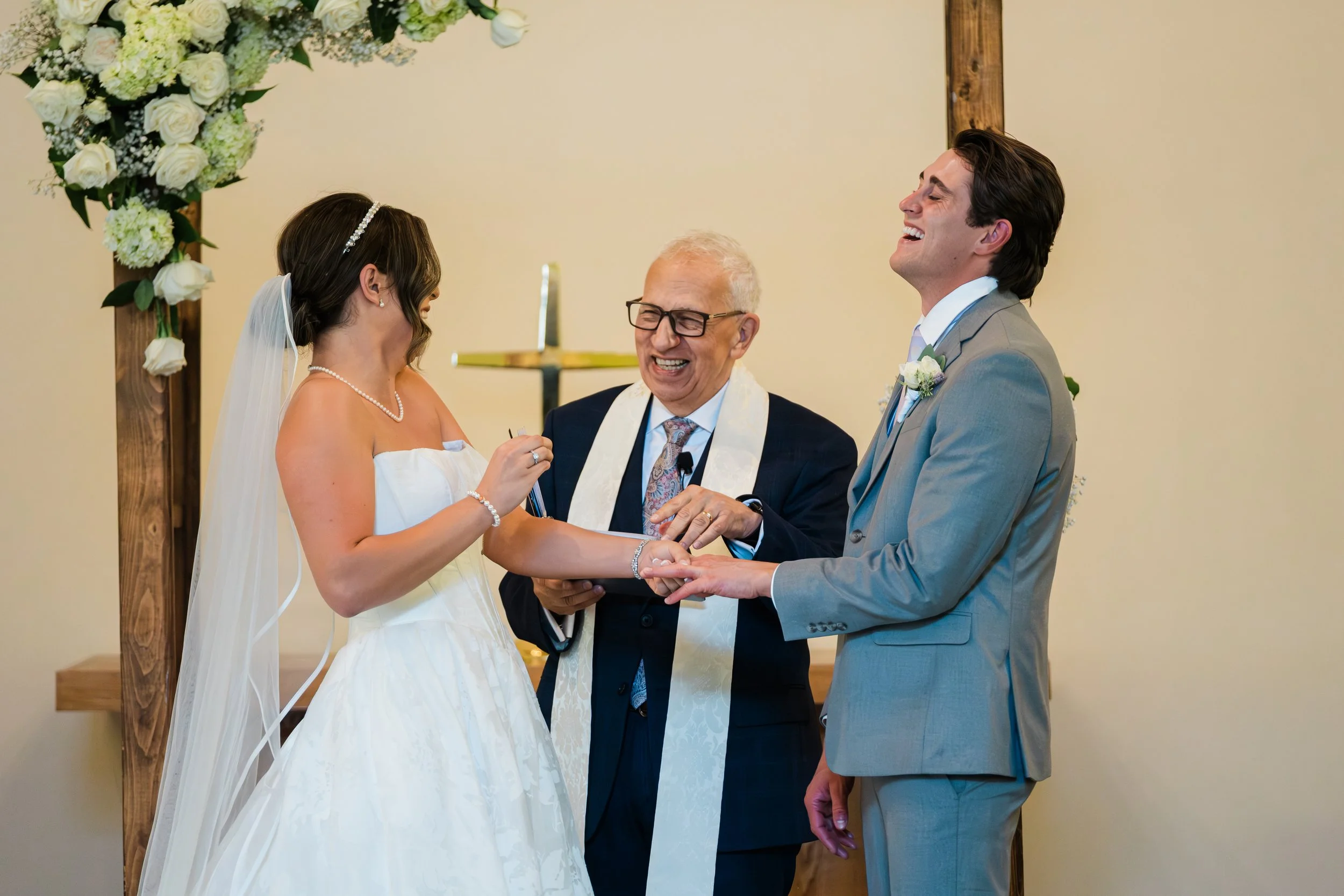 A bride and groom exchanging rings during a wedding ceremony with a officiant, in front of a floral arch.
