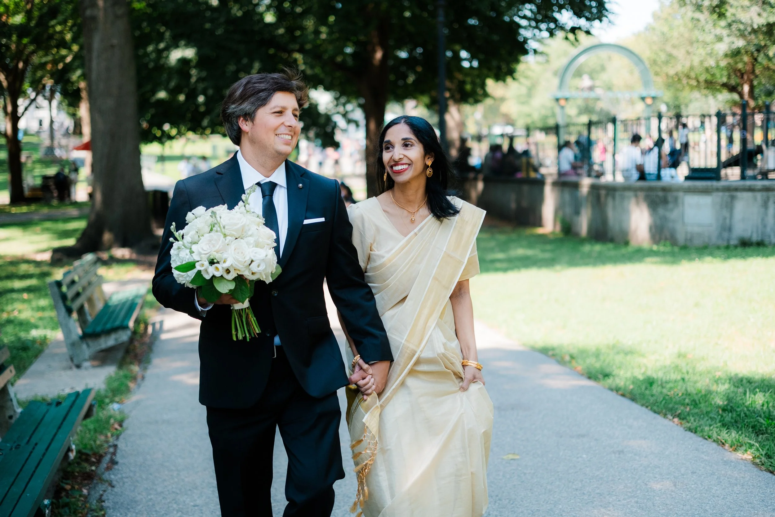 A couple walking hand in hand in a park, dressed formal. The man in a black suit holding a bouquet of white flowers, and the woman in a traditional cream-colored saree, smiling and looking at each other.