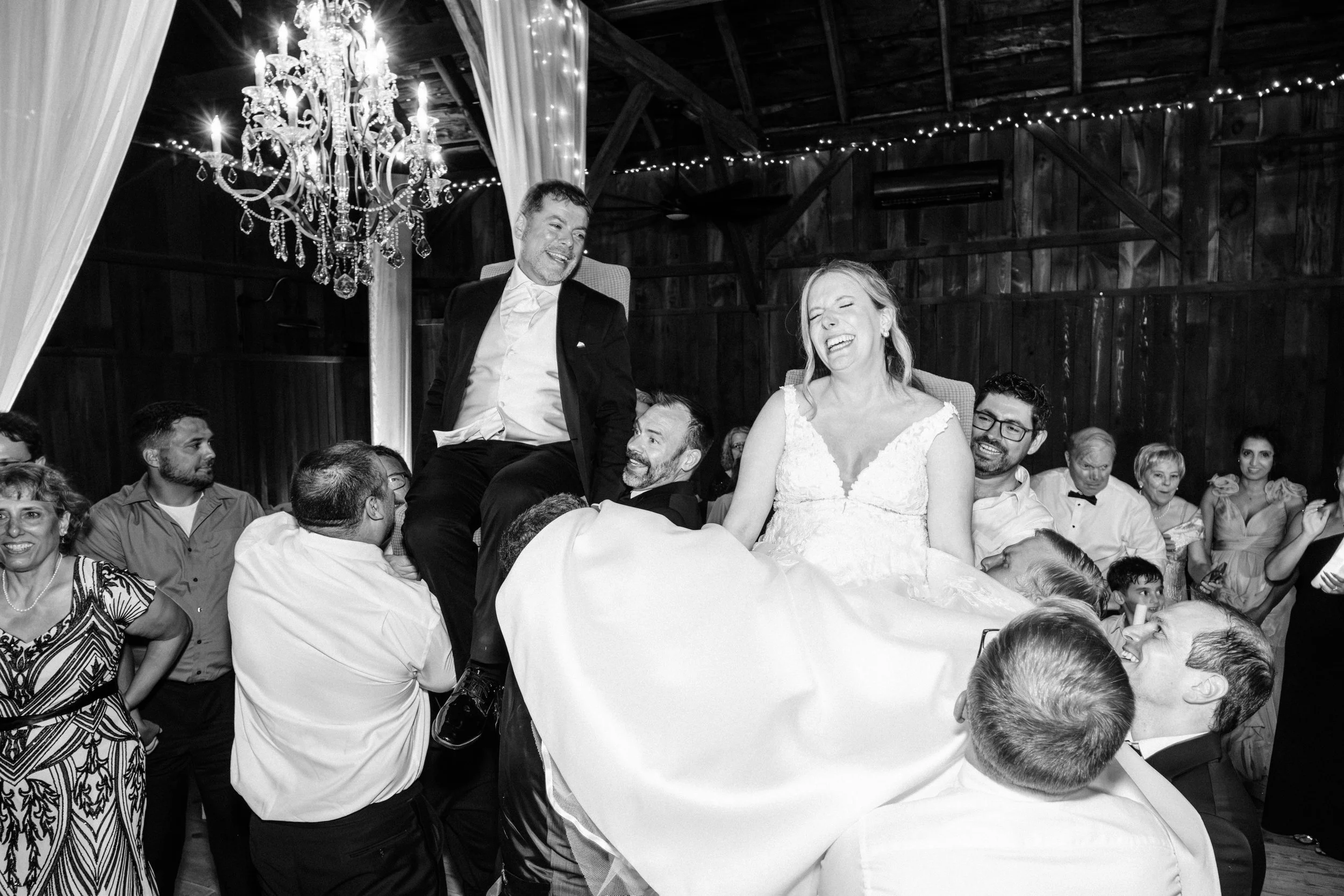 A black and white photo of a groom and bride laugh during the Hora, a traditional Jewish dance, during a wedding reception in a barn.