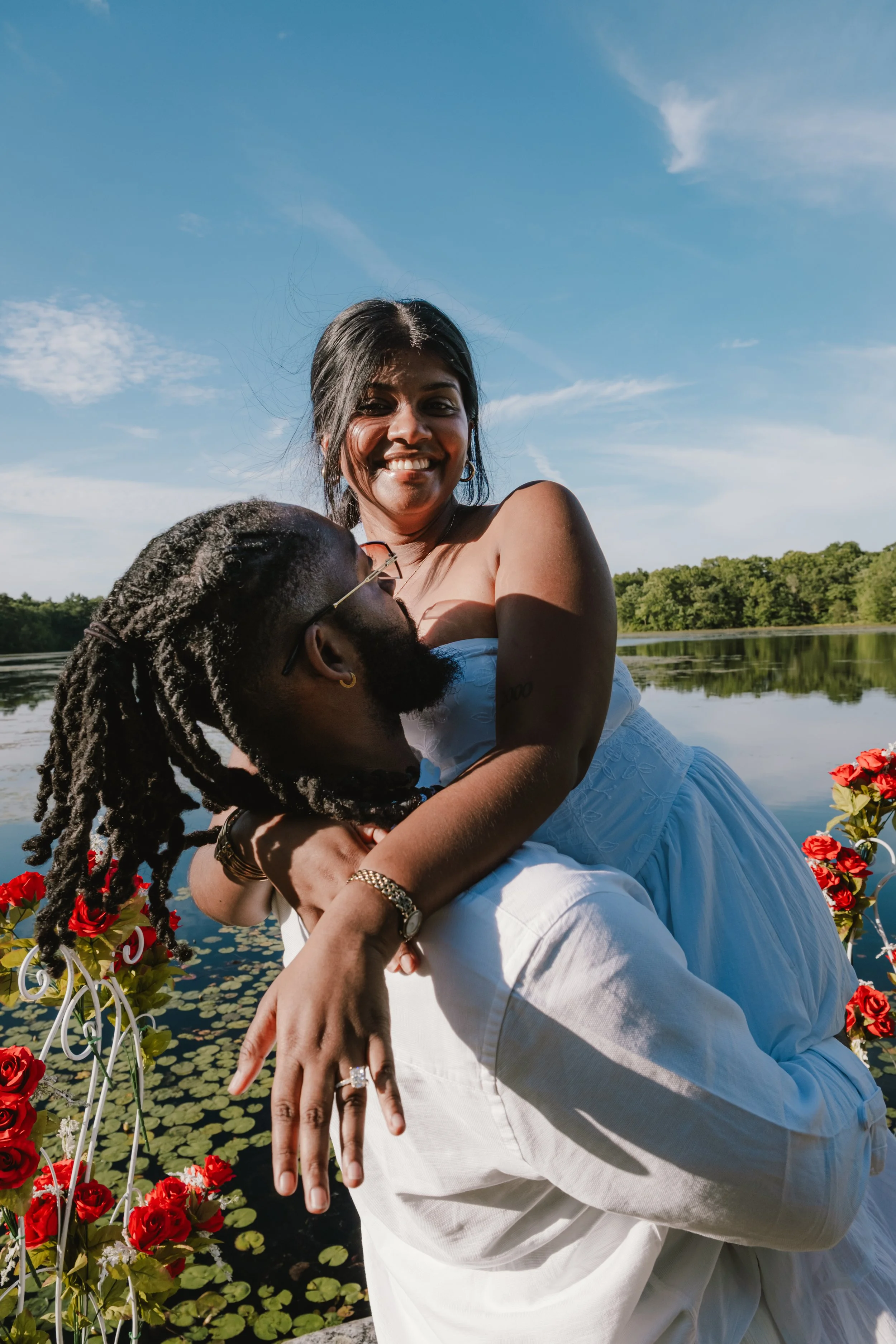 A couple is near a pond with water lilies, the woman is smiling and sitting on the man's lap, surrounded by red flowers, with a background of trees and a blue sky.