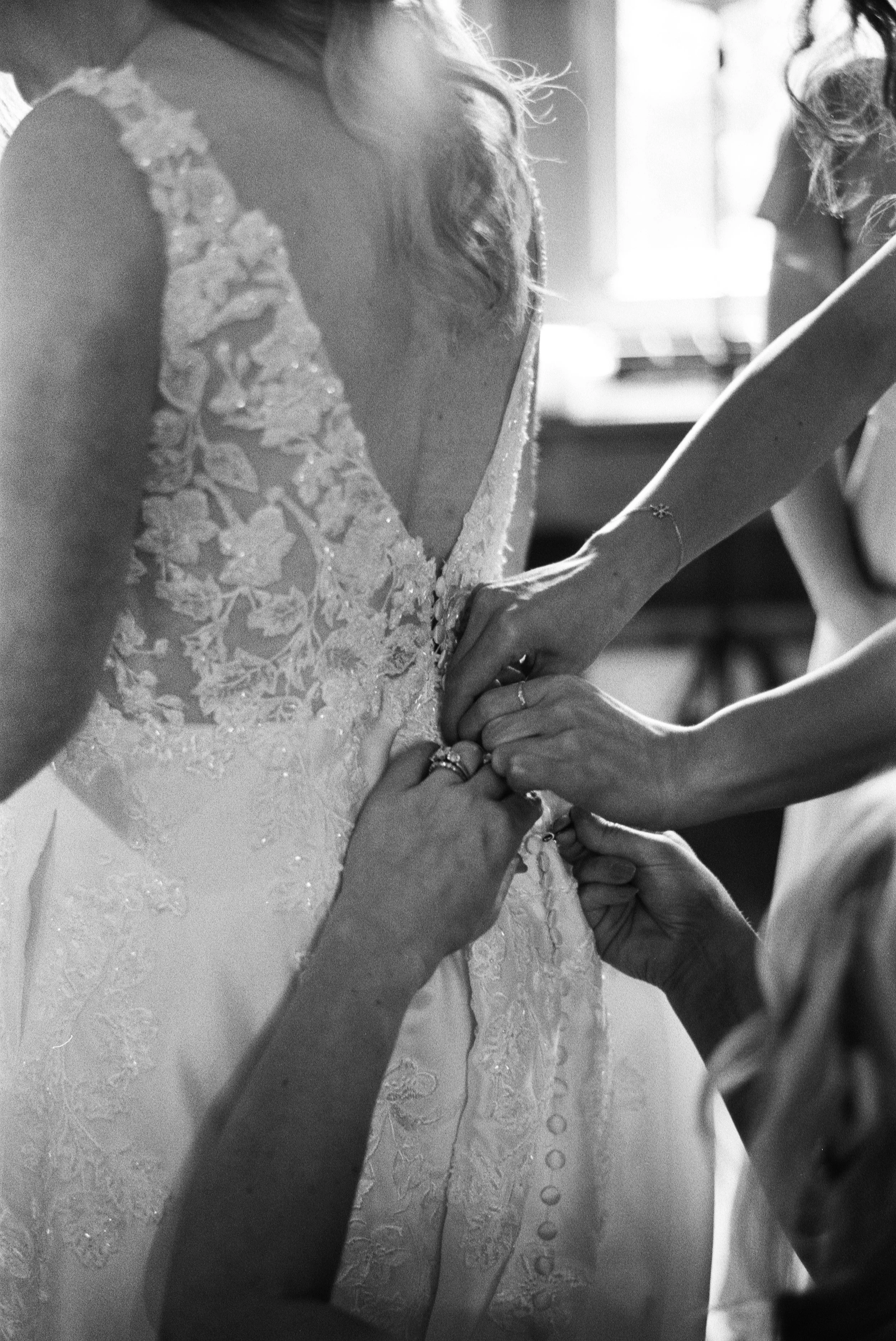 Black and white film photo of a bride getting her wedding dress buttoned up by bridesmaids while getting ready for her Maine wedding.