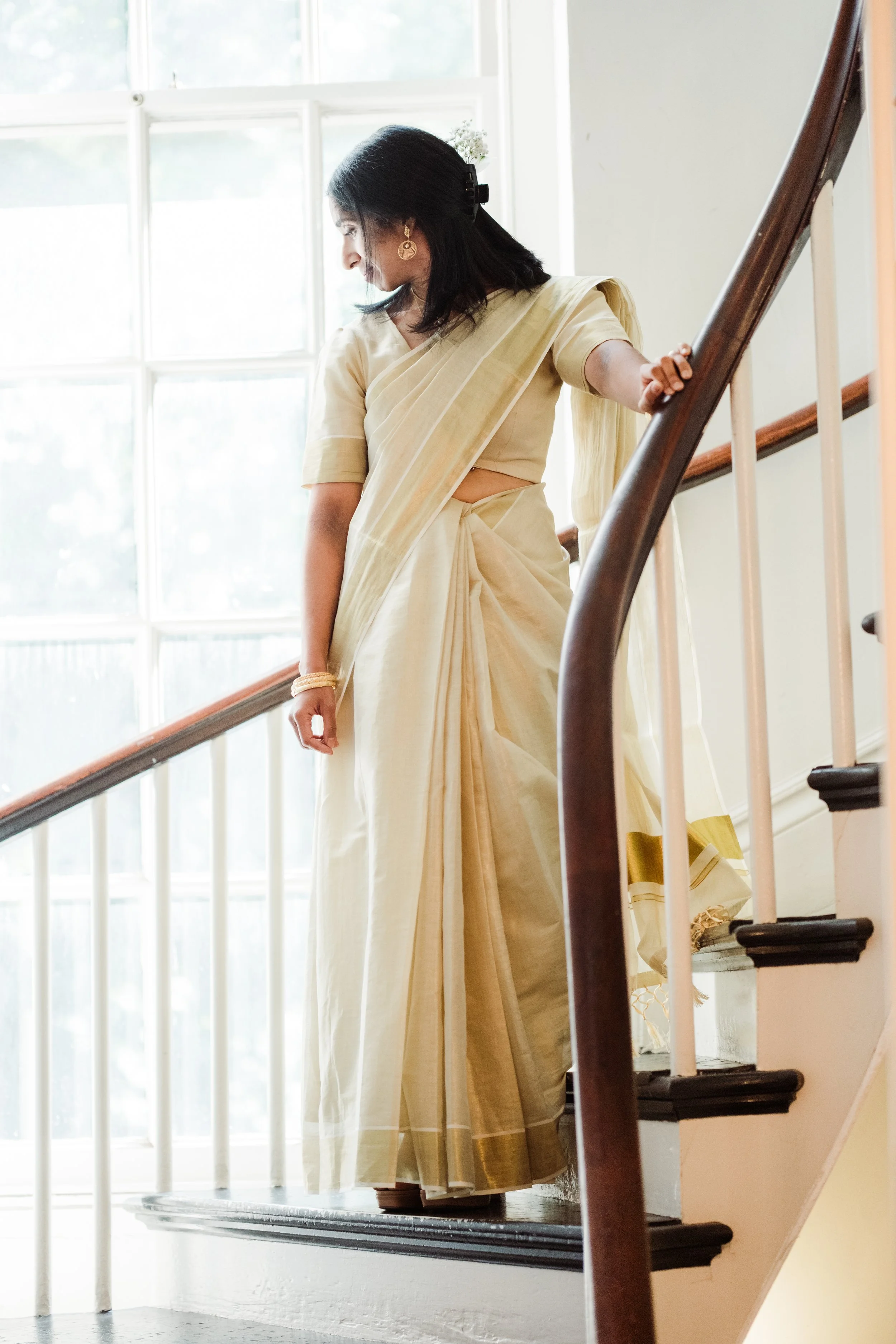 A woman in a cream-colored saree standing on a staircase by a large window, looking down.