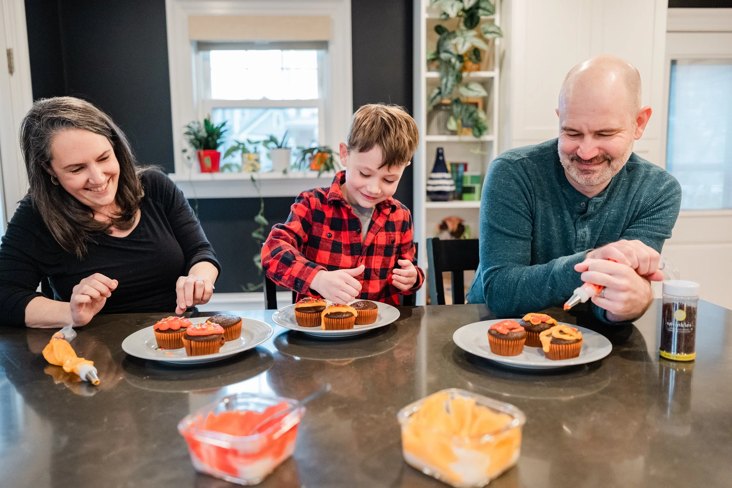 Family decorating cupcakes at a kitchen table, with three people smiling and applying orange and pink frosting to cupcakes.