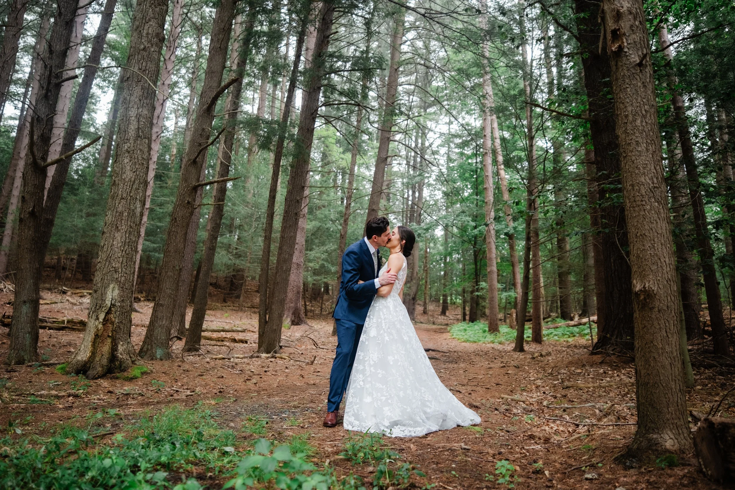 A couple in wedding attire kissing in a forest surrounded by tall trees.