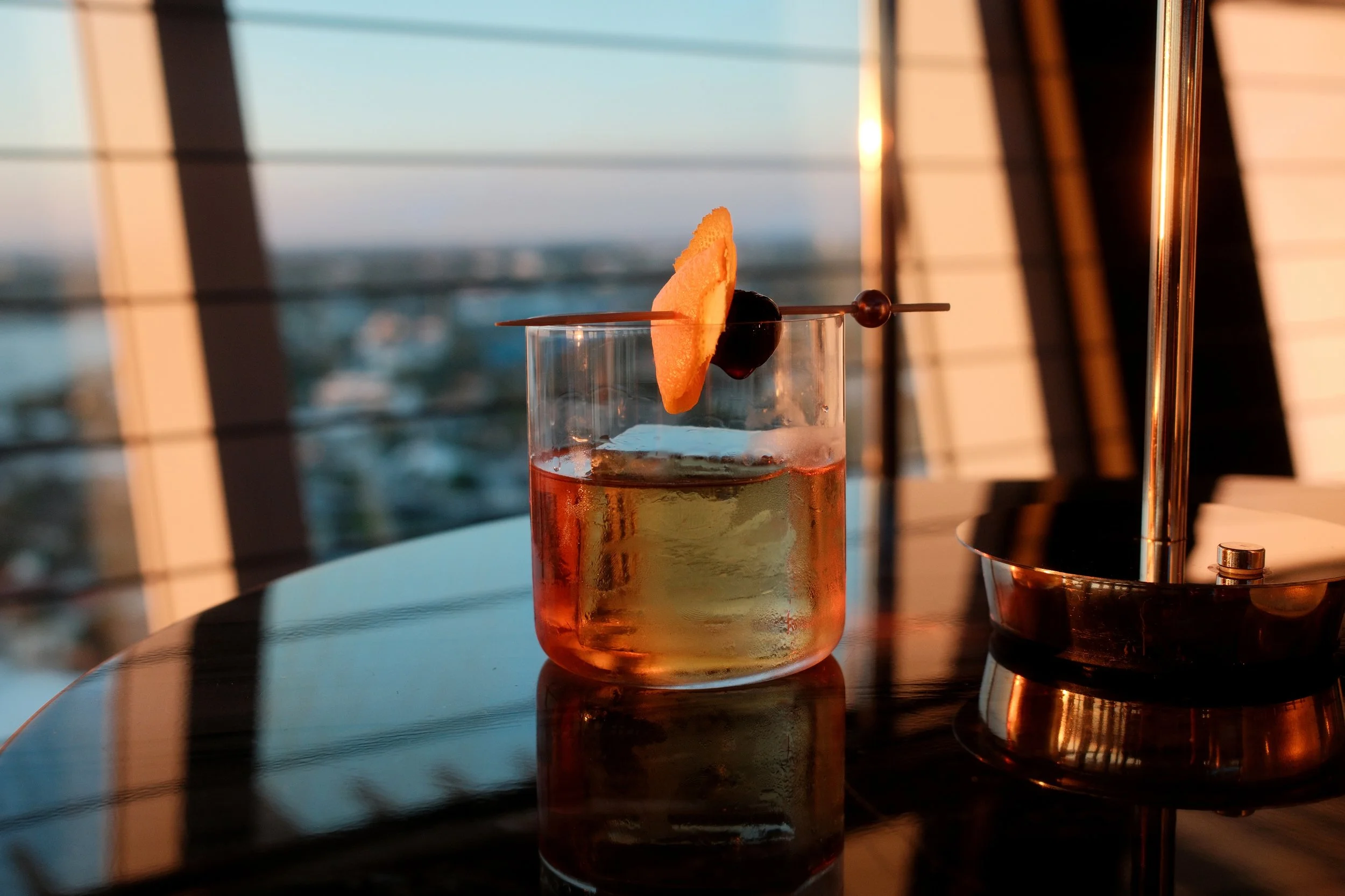 A cocktail, garnished with an orange slice and a cherry, sitting on a reflective table near a sunset window at Pier Sixty-Six in Fort Lauderdale, Florida