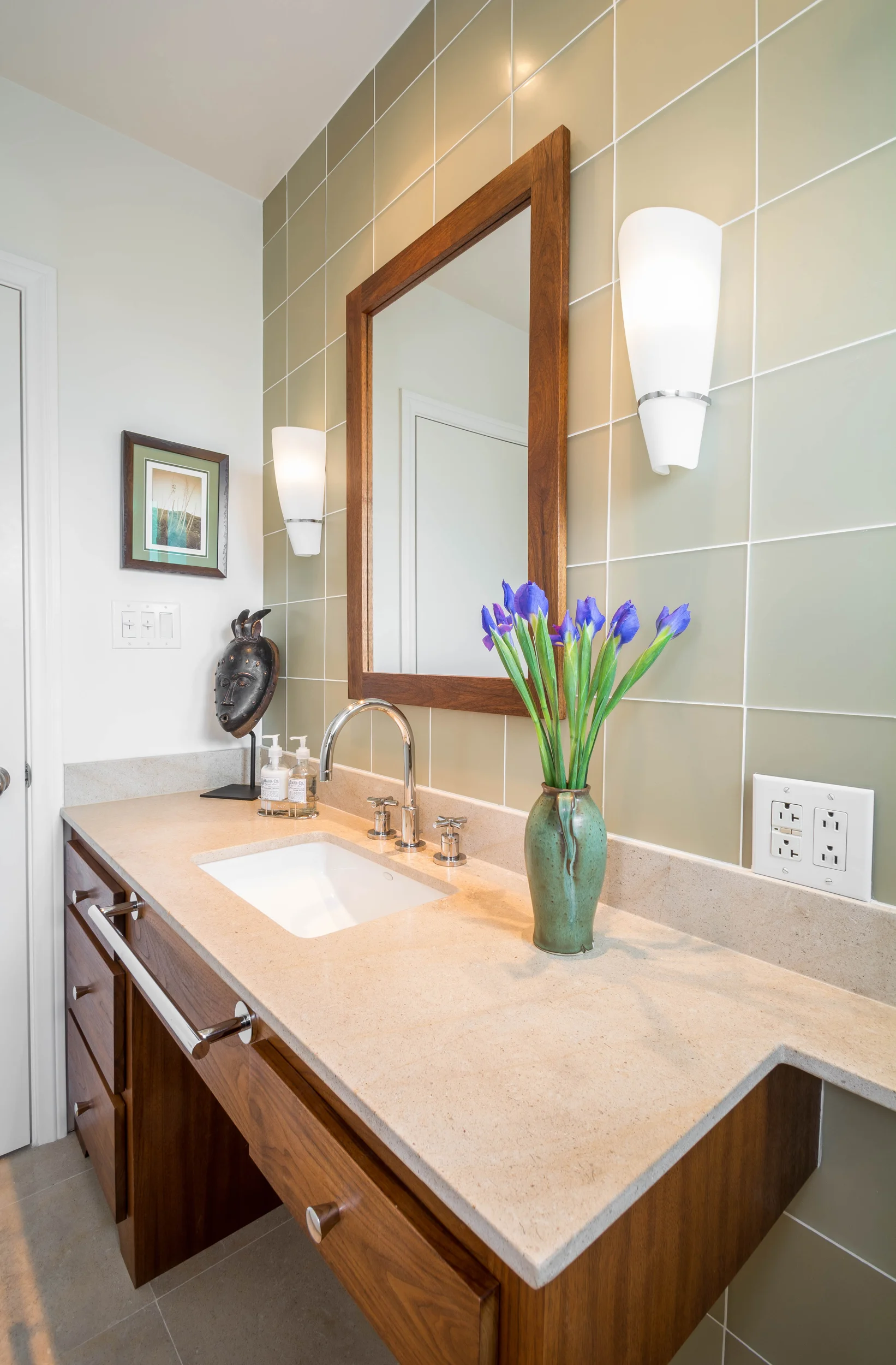 This sophisticated powder room features a custom-built walnut mirror frame and bathroom vanity accentuated by a limestone top, matte glass tiles and modern sconce light fixtures
