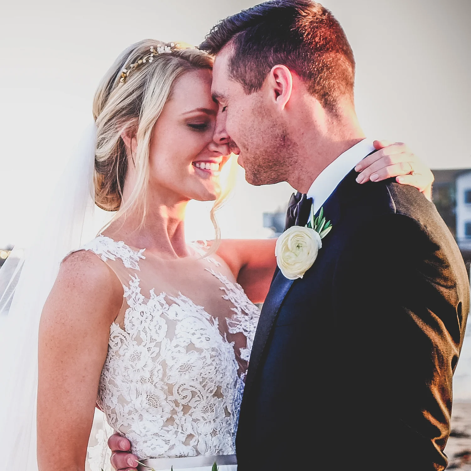 A bride and groom with foreheads touching, smiling at each other, outdoors during sunset, with a blurred background.