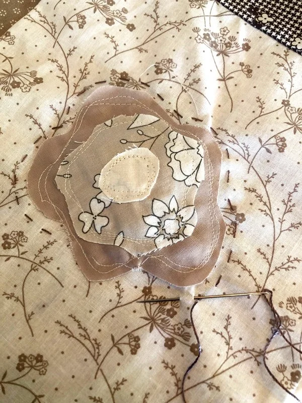 A needle is stuck into the a floral beige and white fabric. three layers of fabric are sewn on the quilt in a floral shape that suggests a rose. 