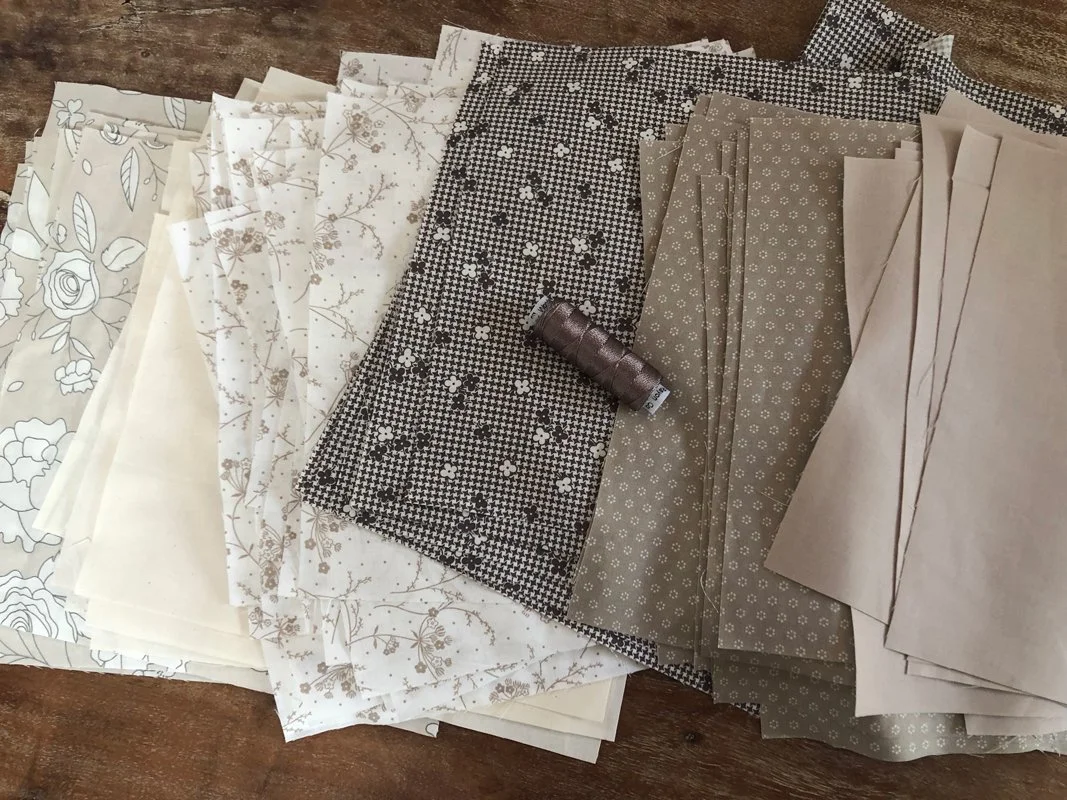 Squares of fabric stacks laying in a gradient of white, cream, beige, khaki, light brown and browns. A medium dark thread is on top. 