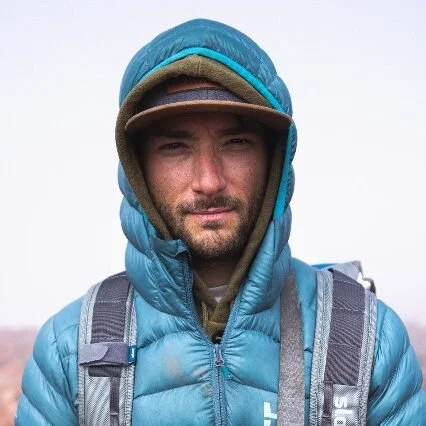 Man wearing a blue hooded jacket with a backpack and a cap underneath the hood.