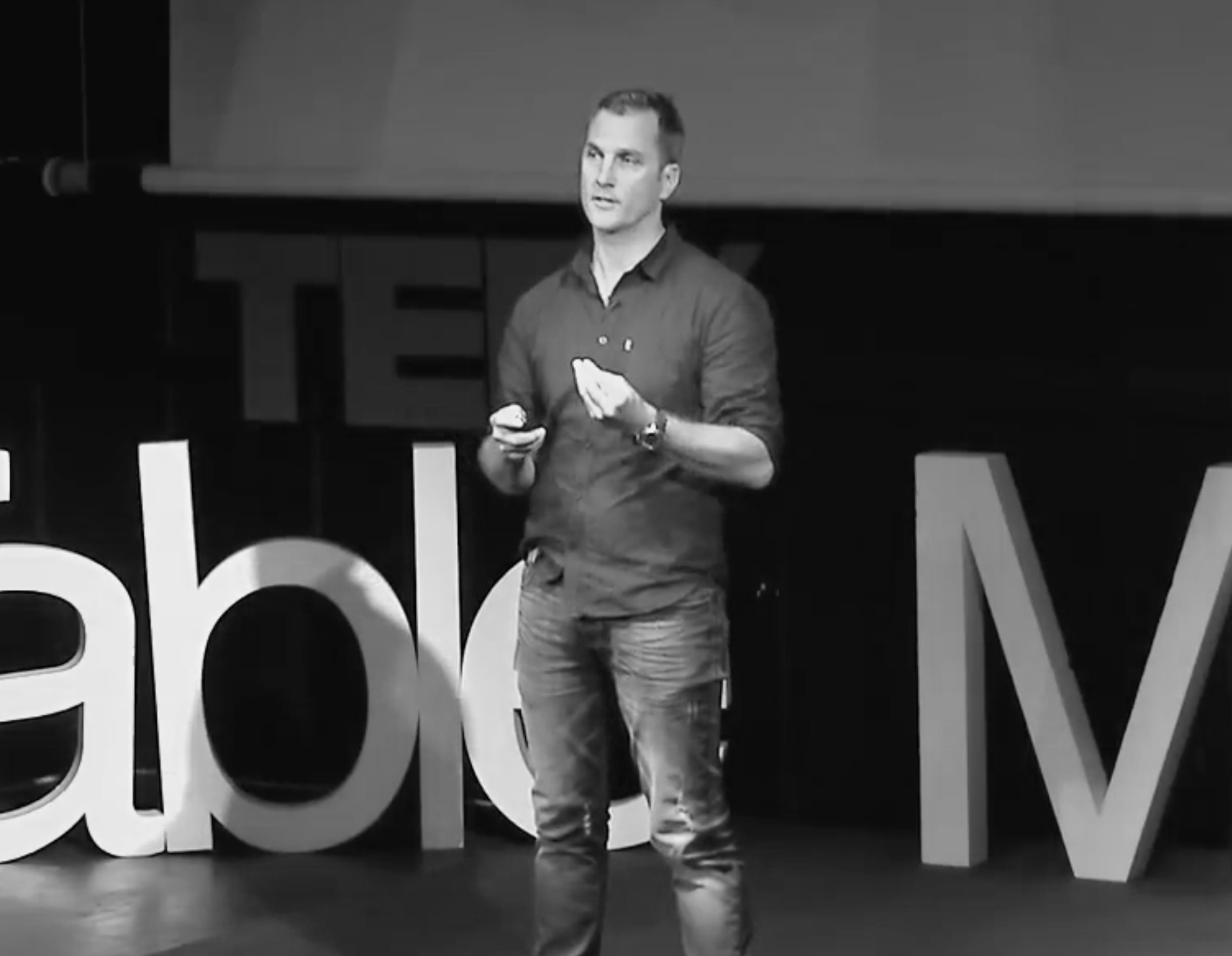 A man with short hair and a watch on his left wrist, wearing a dark shirt and jeans, is speaking on stage with a monitor in his right hand. He stands in front of a large sign with partially visible letters. The scene is in black and white.