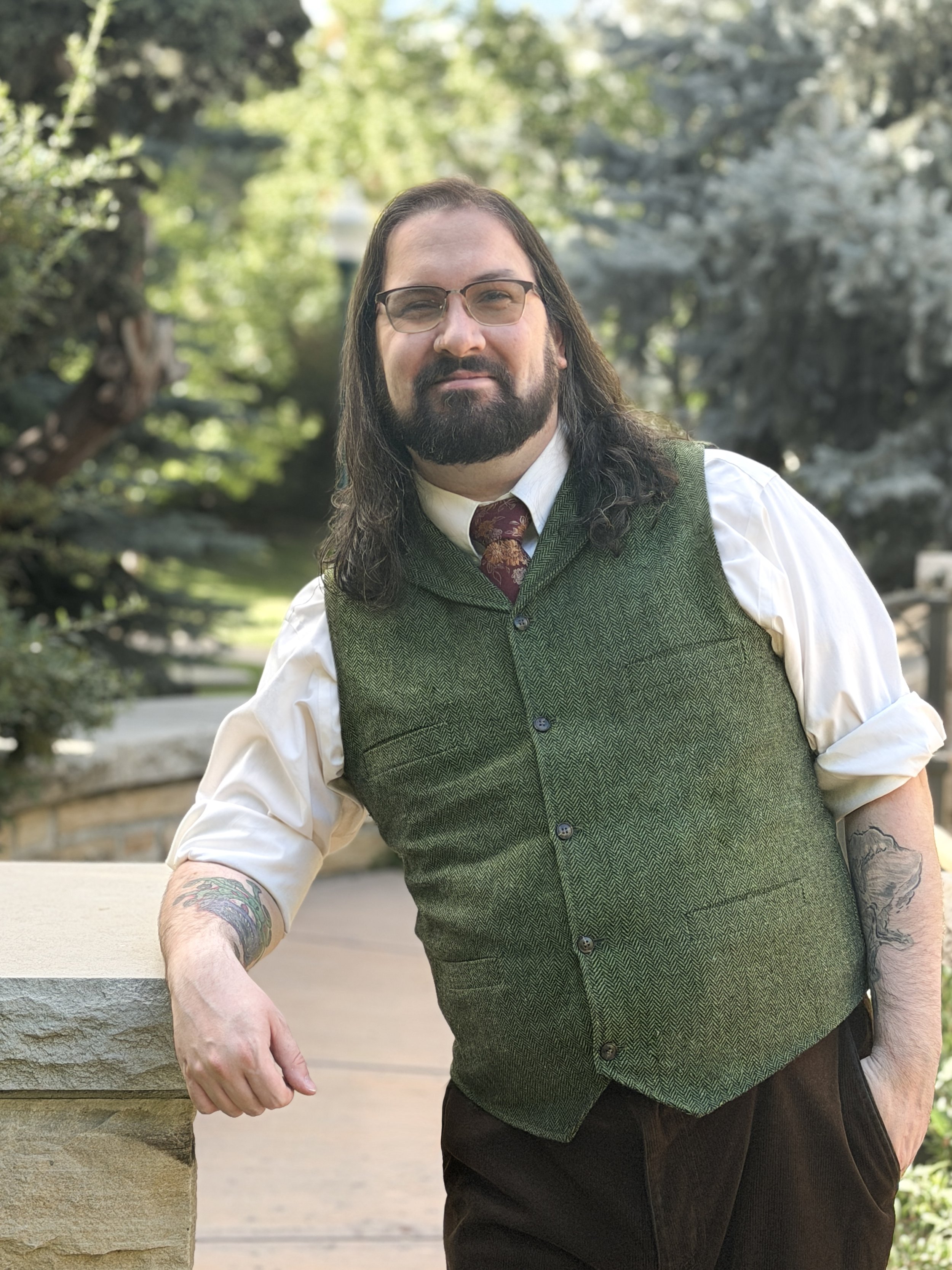 A man with glasses and long dark hair, wearing a green vest over a white shirt and a patterned tie, leaning against a stone wall outdoors with trees in the background.