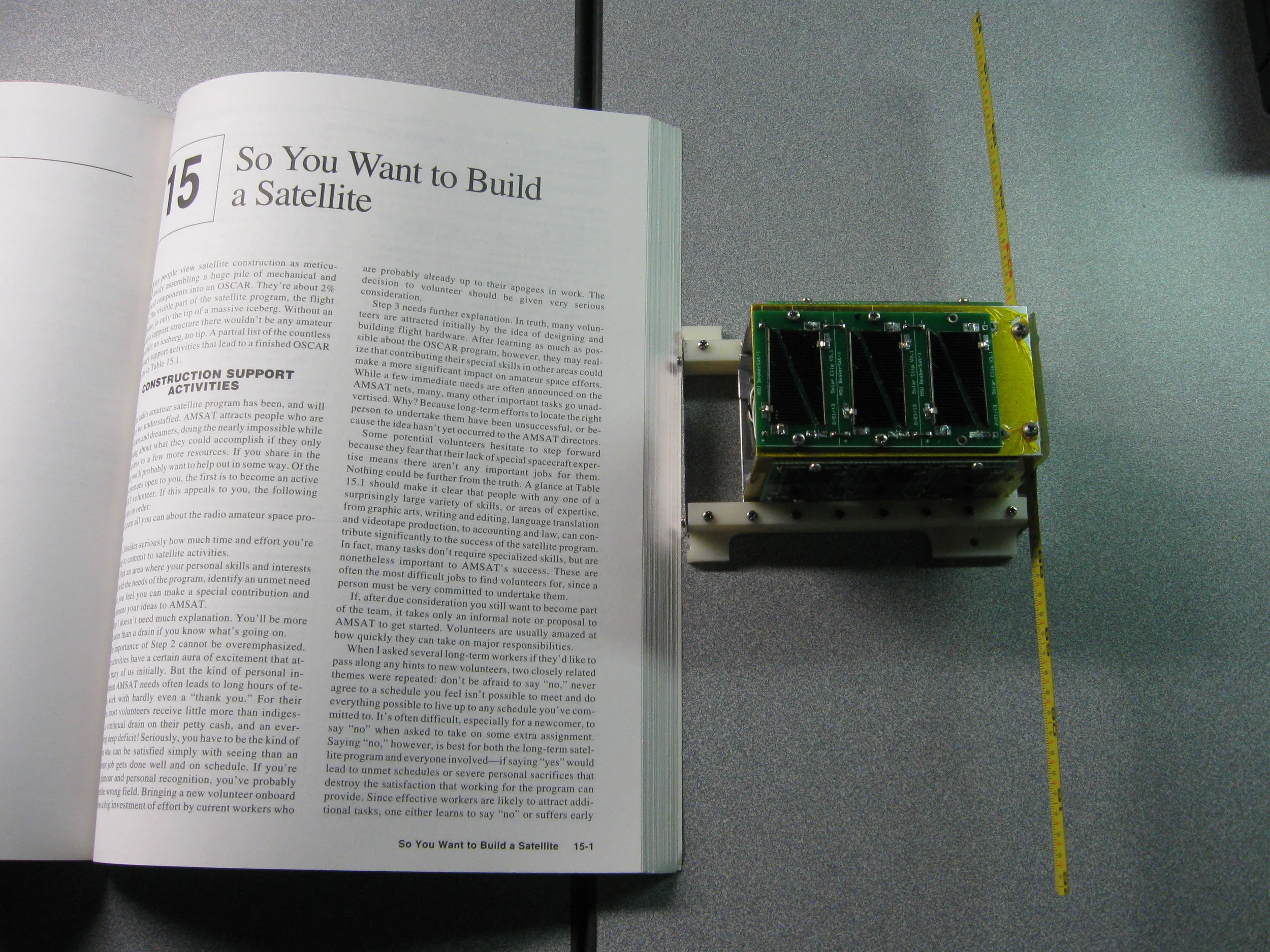 The $50sat PocketQub shown next to an inspirational page for scale. Take a look at the satellite's yellow antennas. Just like the original Oscar, the budget satellite uses a metal tape measure rather than an expensive high-tech antenna. Source: 50dollarsat.info