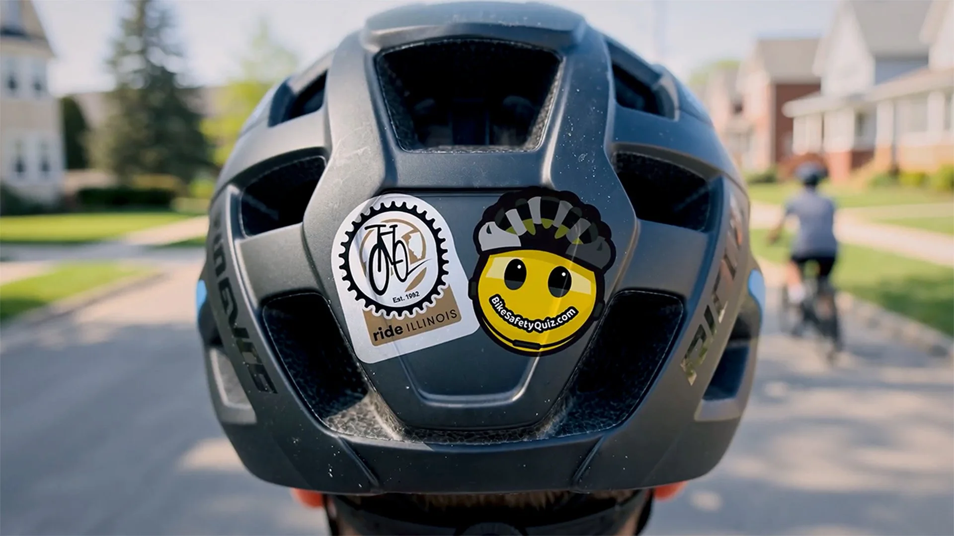 Backside of a black bike helmet on a bicyclist riding through a suburban IL neighborhood. Two stickers featuring the RideIllinois.org logo and BikeSafetyQuiz.com smiley face are stuck to the back of the helmet