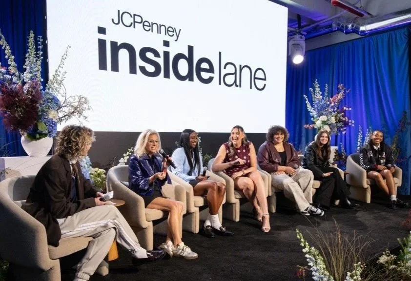 Panel of seven women sitting on chairs on stage at an event for JCPenney Inside Lane, with a large screen behind them displaying the event name and floral decorations on either side of the stage.