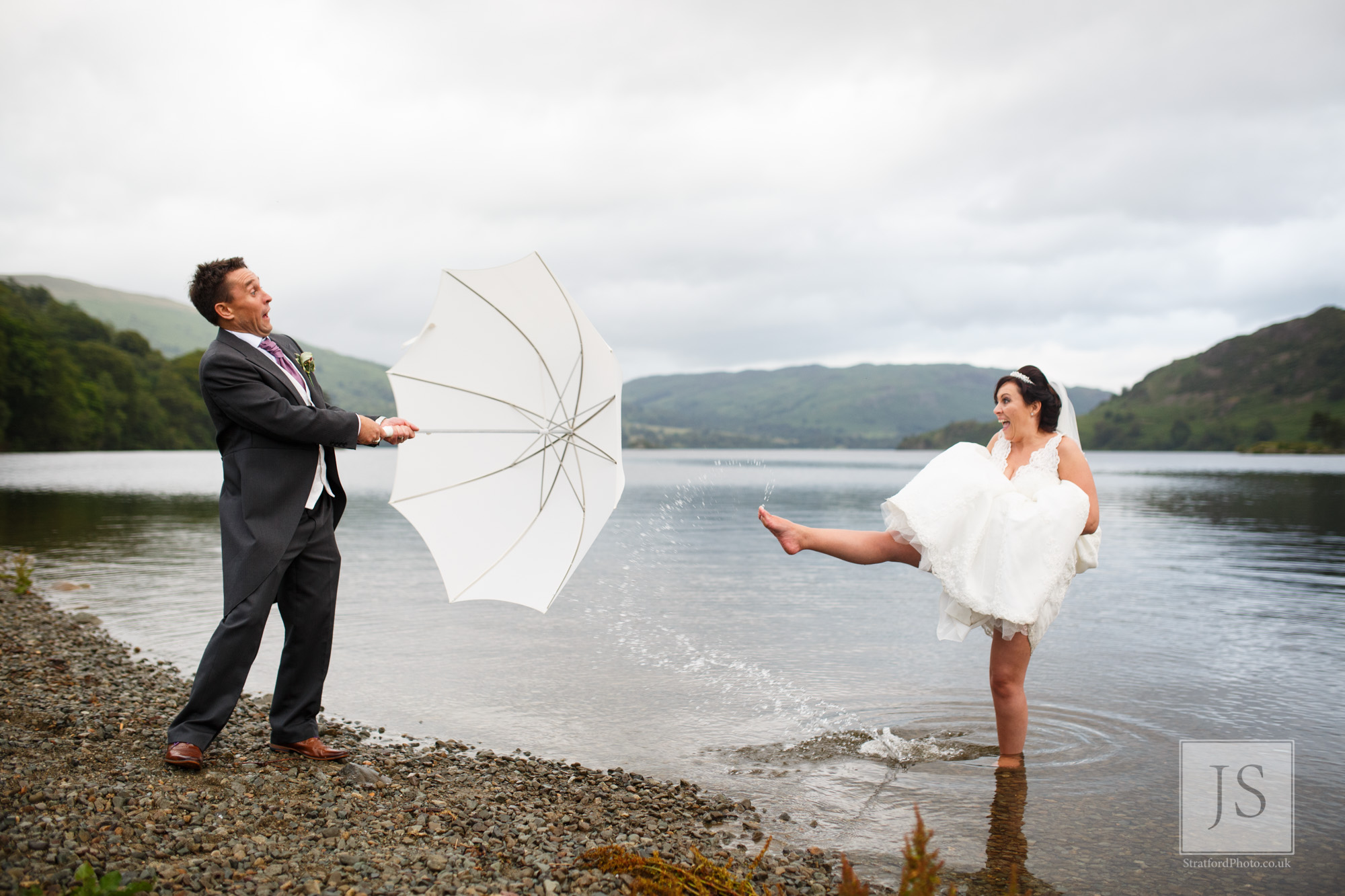 A bride kicks Lake Ullswater water at her groom as he holds up an umbrella.jpg