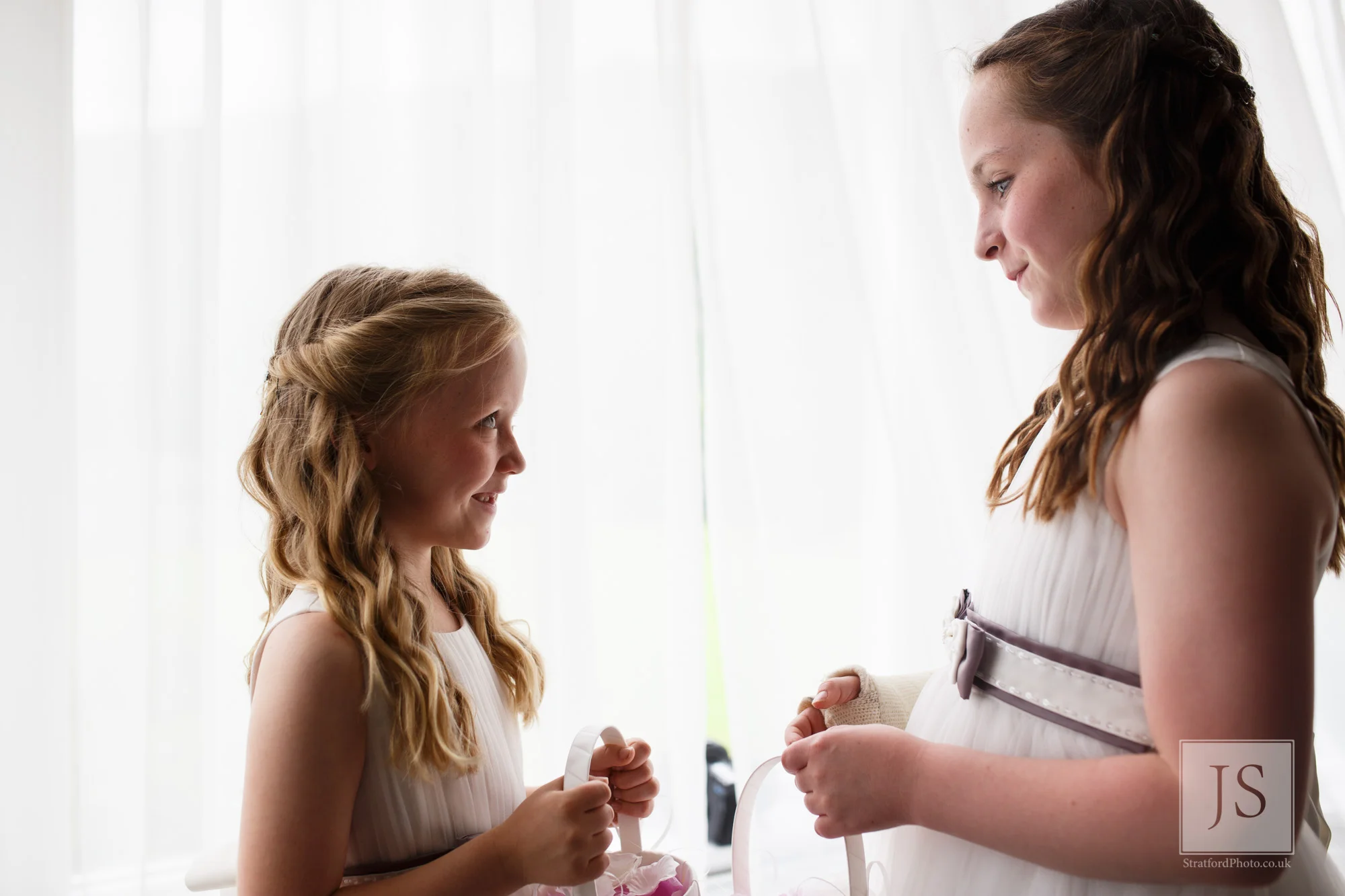 Two beautiful flower girls smile at each other in window light.jpg