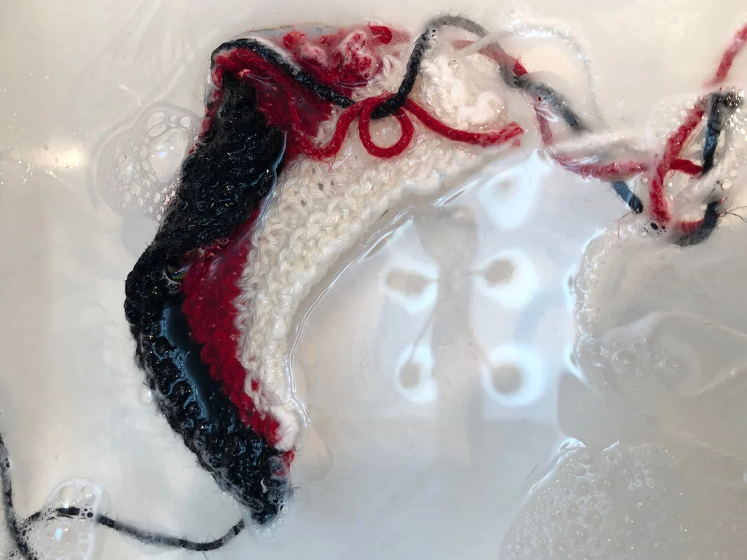 A close up view of a red, white, and black knitted swatch soaking in the sink. Sideways Swatching || withwool.com #knitswatch #darkmatterknits