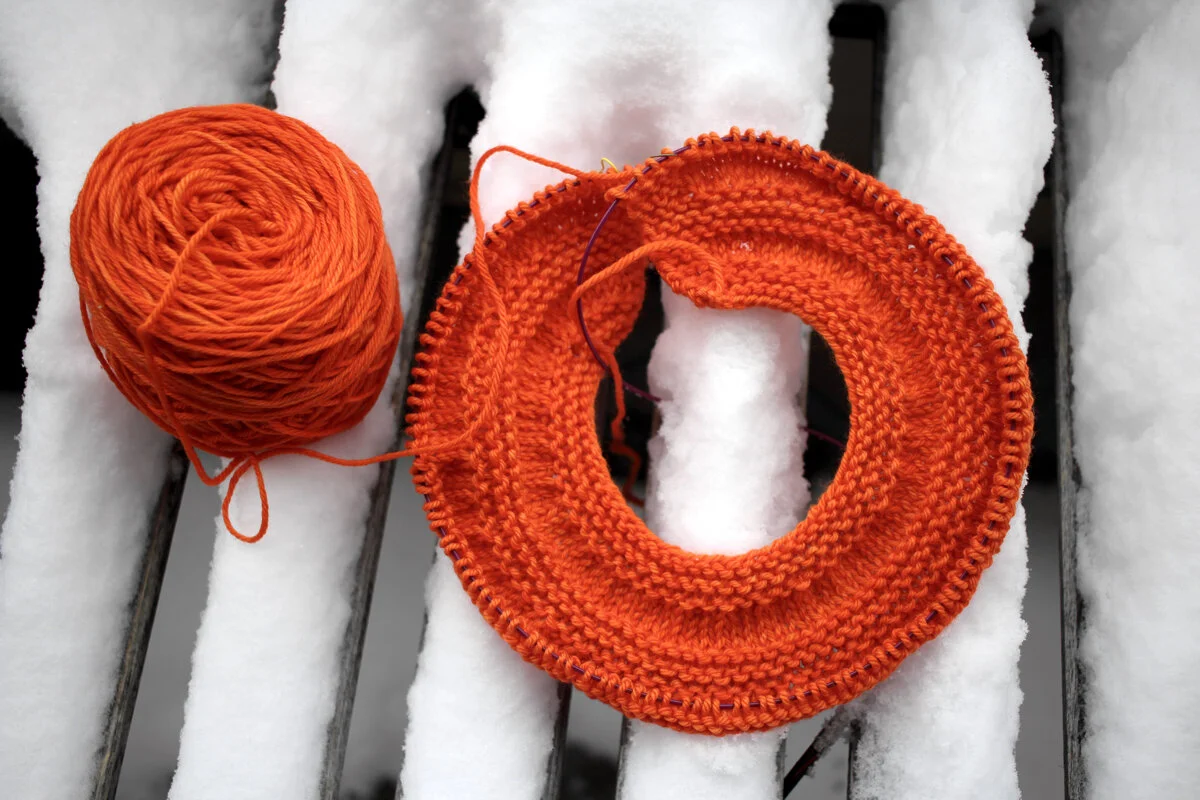 An orange ball of yarn and an in progress sweater yoke sit on a table covered in snow. #knitting Sweater Weather || withwool.com