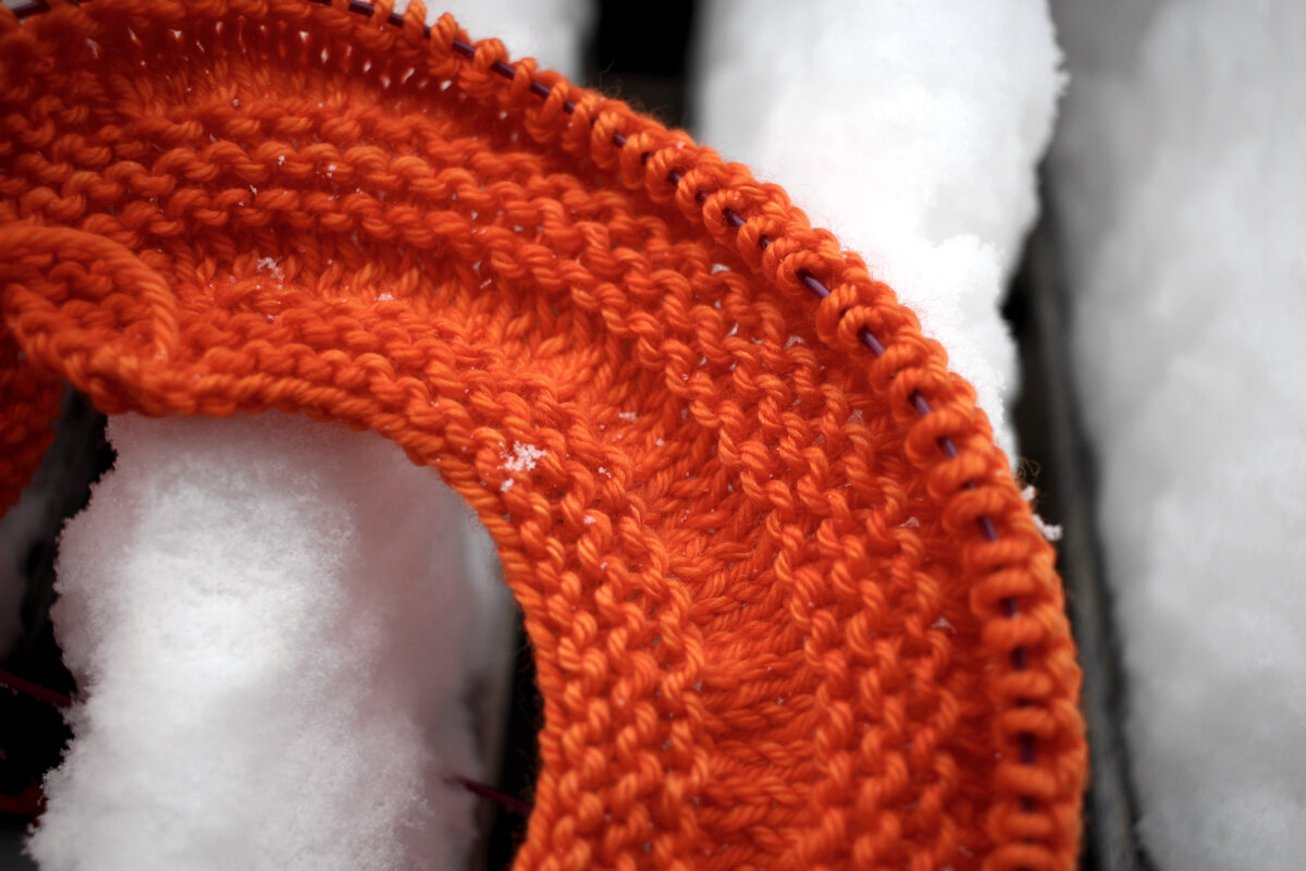 An orange ball of yarn and an in progress sweater yoke sit on a table covered in snow. #knitting Sweater Weather || withwool.com