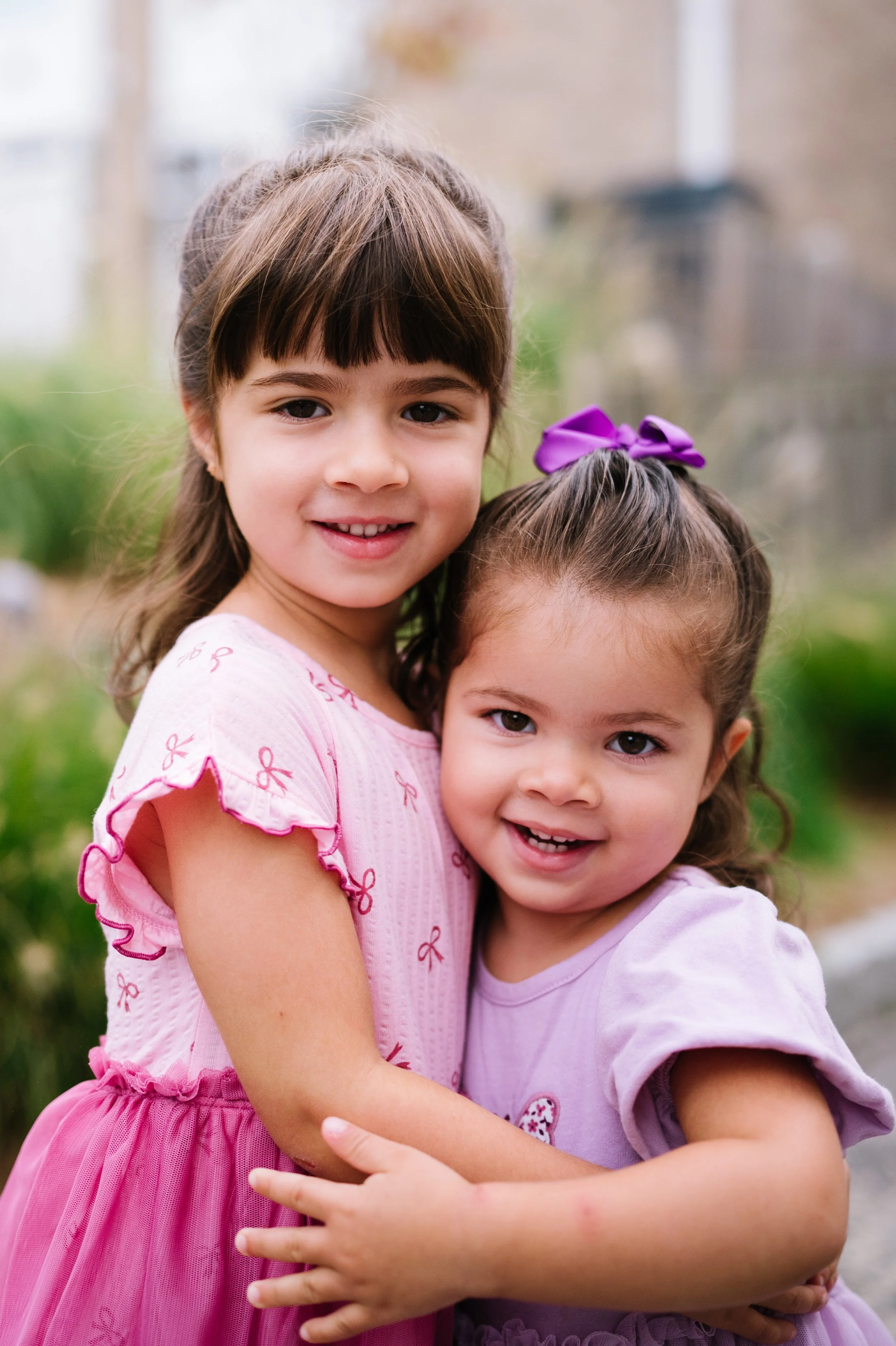 Two young girls hugging and smiling outdoors, one wearing a pink dress with ruffle sleeves and the other in a purple dress with a purple bow in her hair.