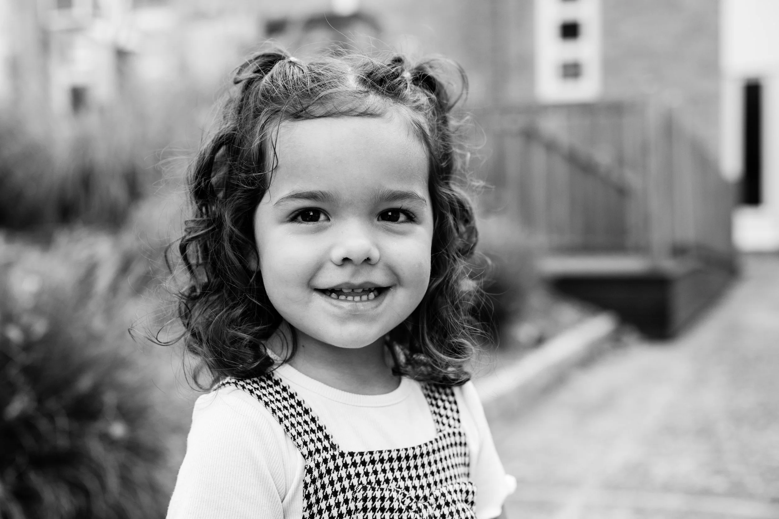 A young girl with curly hair smiling outdoors in black and white.