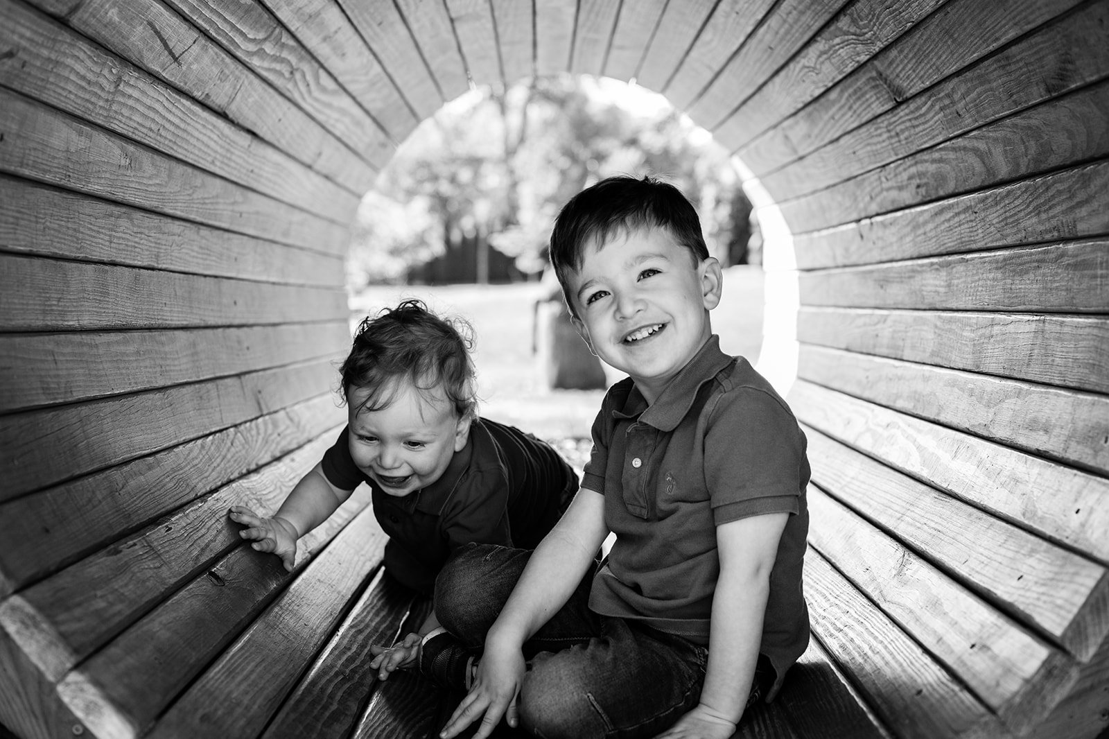 Two young boys playing inside a circular wooden tunnel, smiling and having fun outdoors.