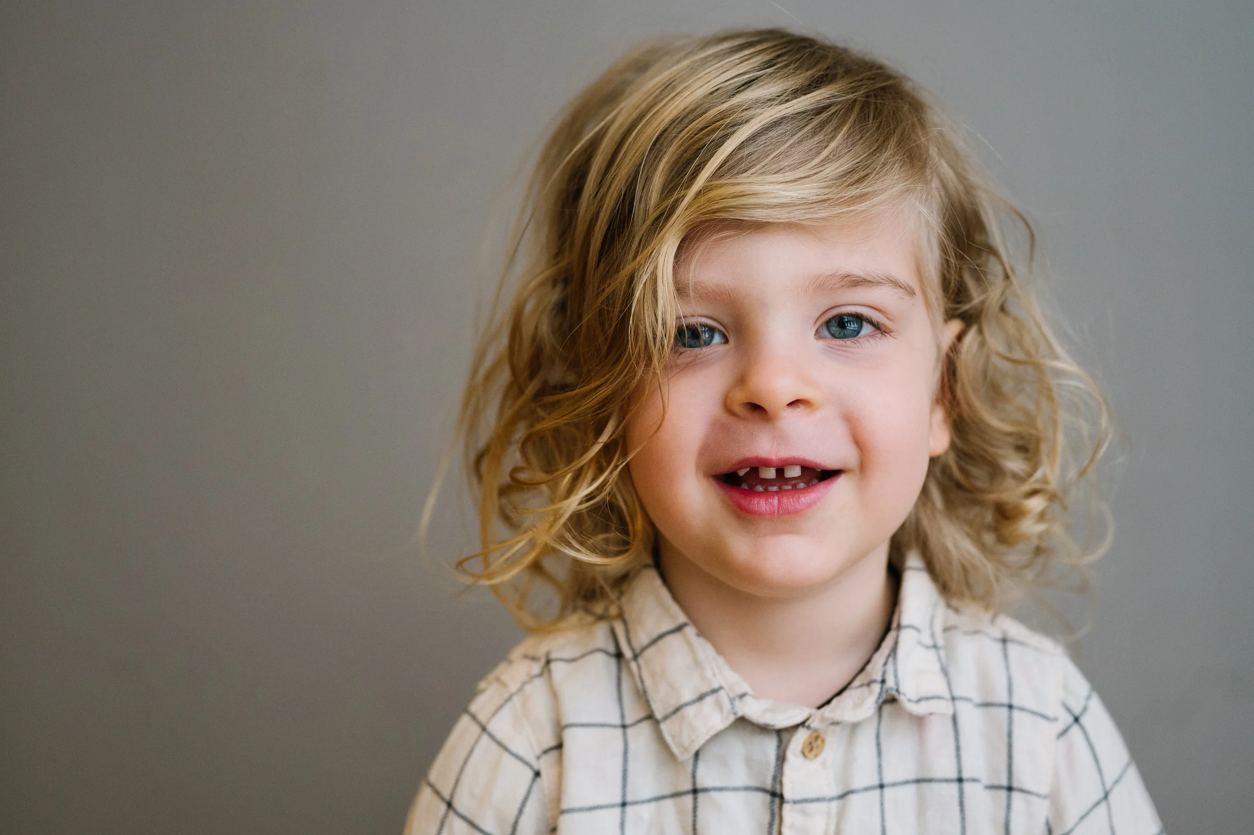 A young child with curly blonde hair, blue eyes, and missing front teeth, smiling and wearing a beige checkered shirt against a plain gray background.