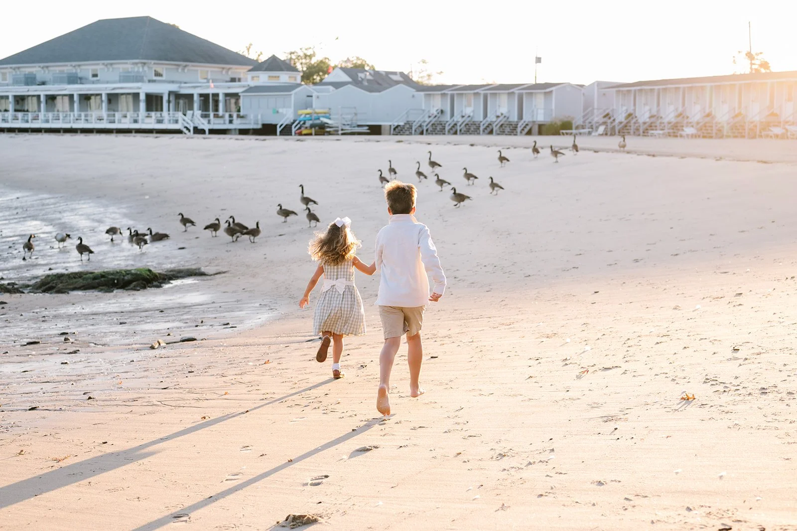 Two children, a girl and a boy, running on the beach at sunset, with gulls on the sand, beachfront houses in the background.