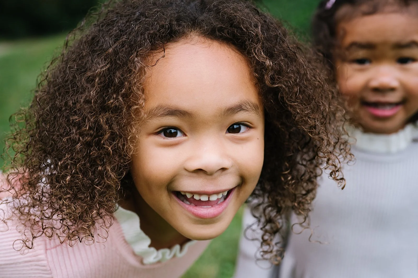 Smiling young girl with curly hair and a girl in the background outdoors.
