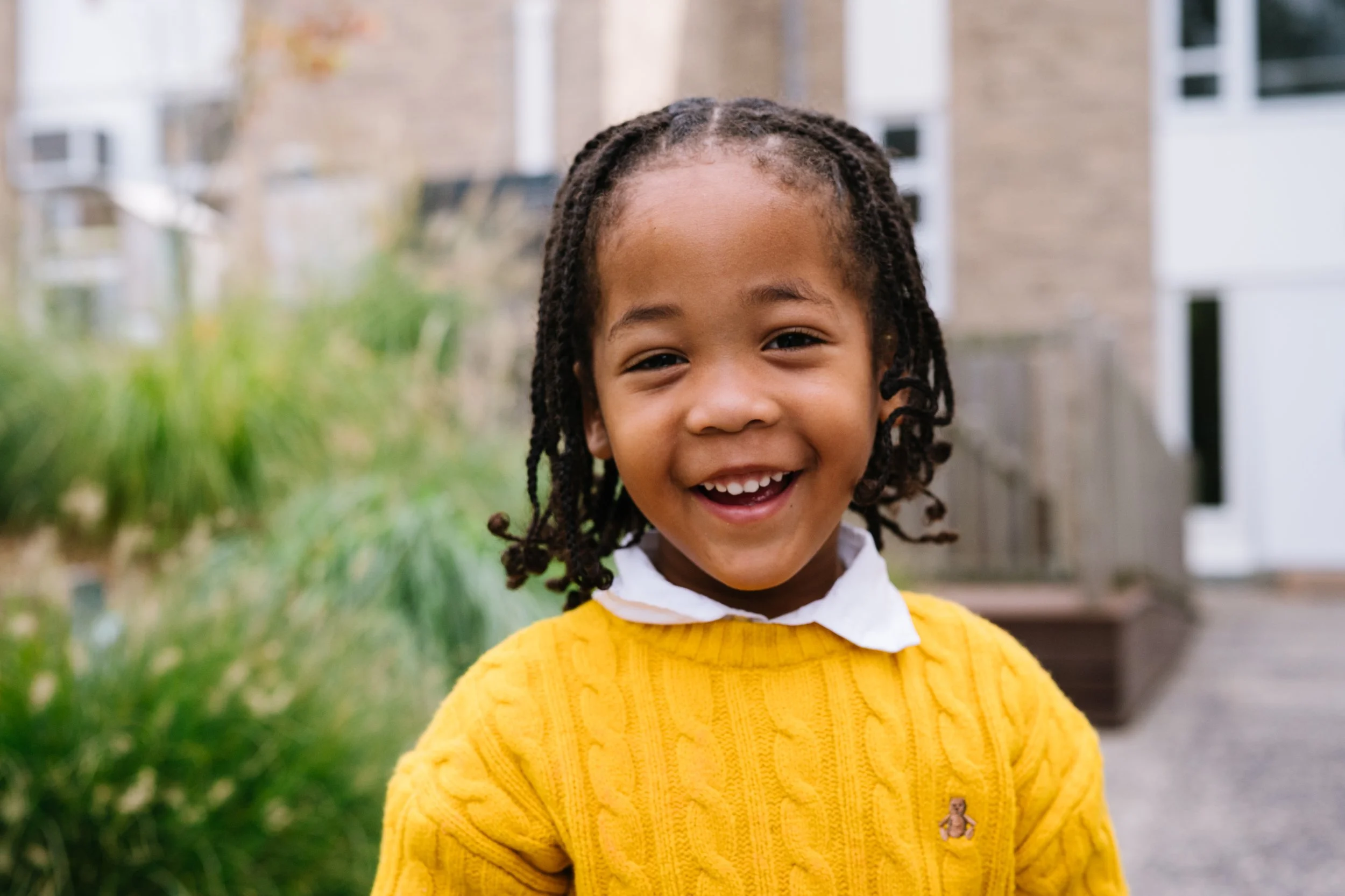 A young girl with braids and a big smile standing outdoors in front of a house.