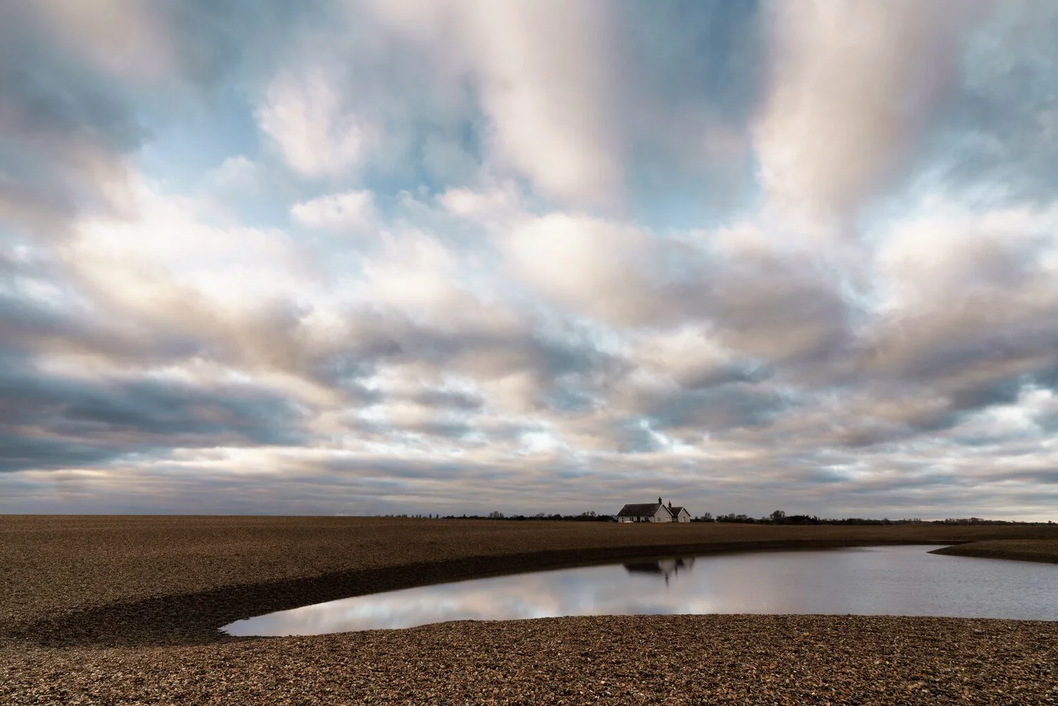 Tidal channel crossing a shingle beach beneath changing clouds
