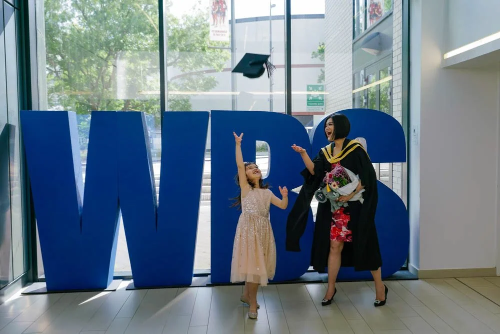 Graduate posing beside large WBS letters inside the university