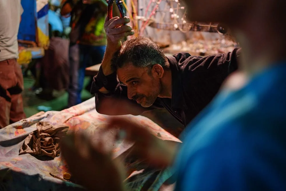 A man taunts young men to shoot the blasting rocks from his hand with air rifles in a carnival game.